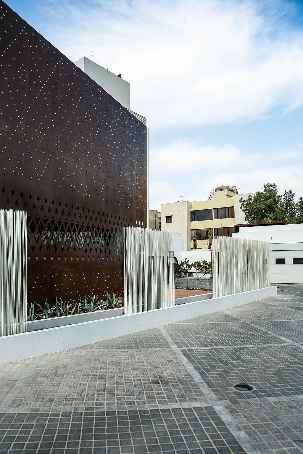 White Sticks rise in separate clusters along the driveway beside the rust-coloured façade, with neighbouring buildings and an open sky in the background.