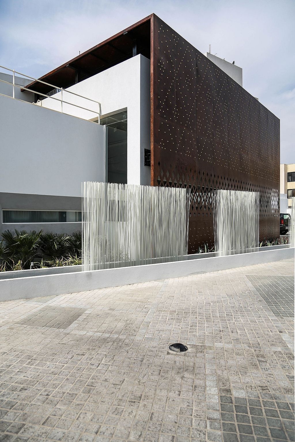 Daytime view of white Sticks arranged in uneven clusters along a low wall beside a white building wrapped by a tall rust-coloured steel screen.