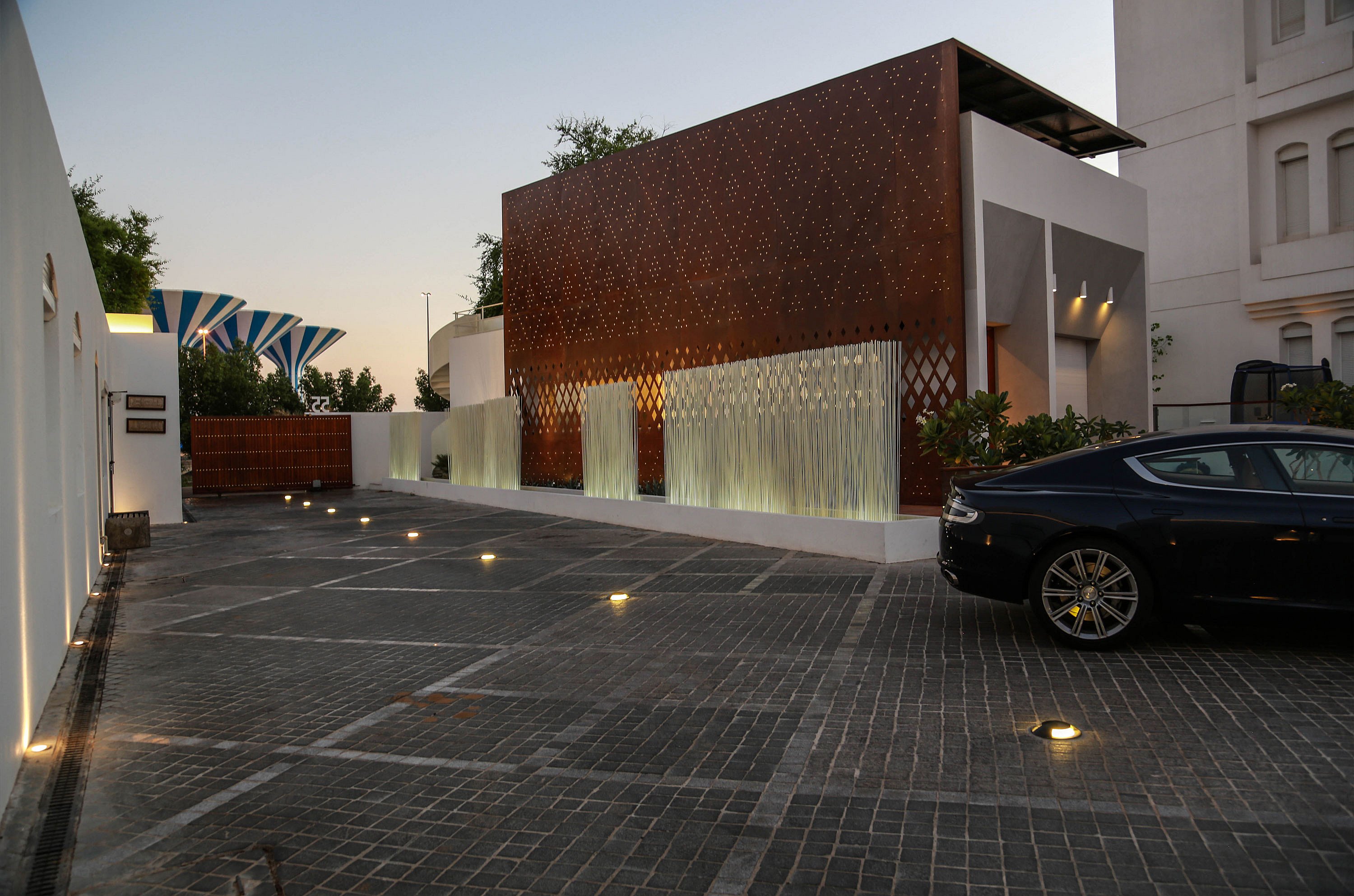 Wide dusk view of the driveway and entrance, where illuminated white Sticks run along the rust-coloured façade and ground lights lead toward a parked car.