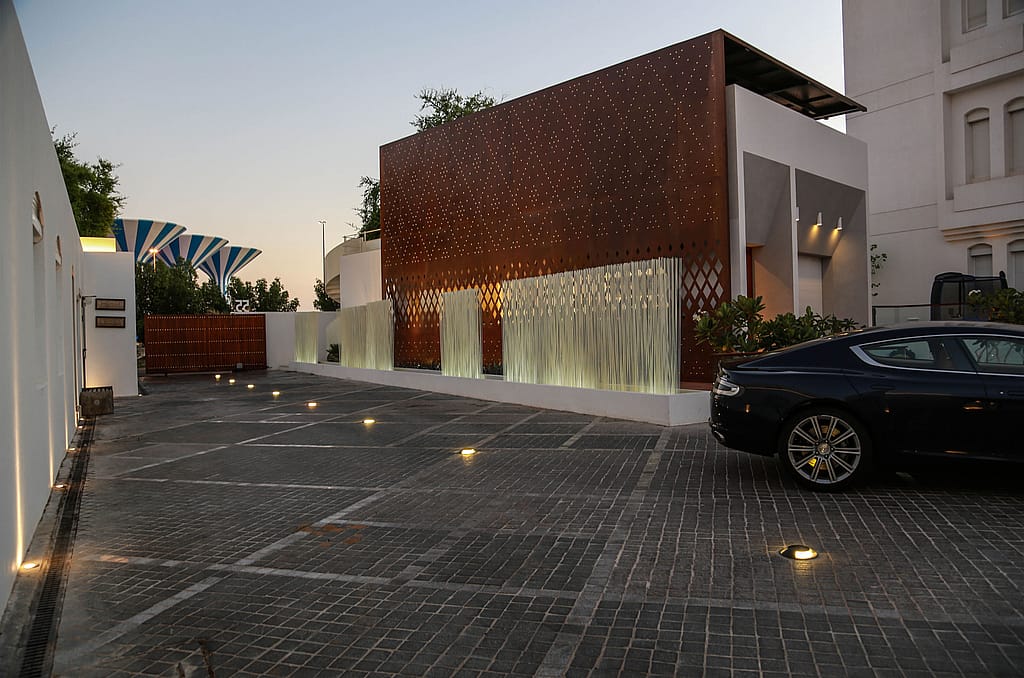 Wide dusk view of the driveway and entrance, where illuminated white Sticks run along the rust-coloured façade and ground lights lead toward a parked car.