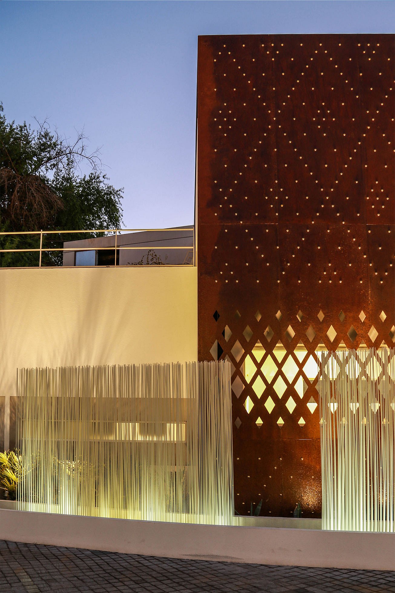 Close-up of white Sticks in front of a rust-coloured steel façade with diamond-shaped cut-outs, highlighting the contrast between the glowing rods and warm metal surface.