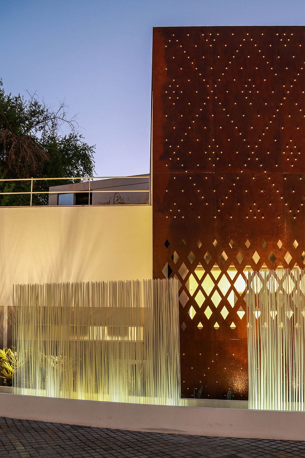 Close-up of white Sticks in front of a rust-coloured steel façade with diamond-shaped cut-outs, highlighting the contrast between the glowing rods and warm metal surface.