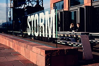 Glaswindschutzscheibe mit dem Wort ‘STORM’ vor der Caféterrasse, wo die Leute an Hopper-Picknicktischen neben dem MAS-Gebäude sitzen.