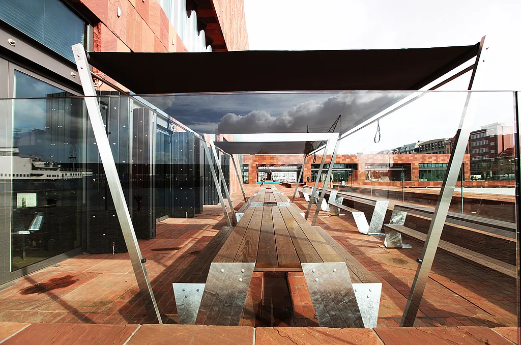A long wood-topped Hopper picnic table stands on the red-stone terrace of Storm Café, framed by glass balustrades and a dark canopy beneath the MAS museum.