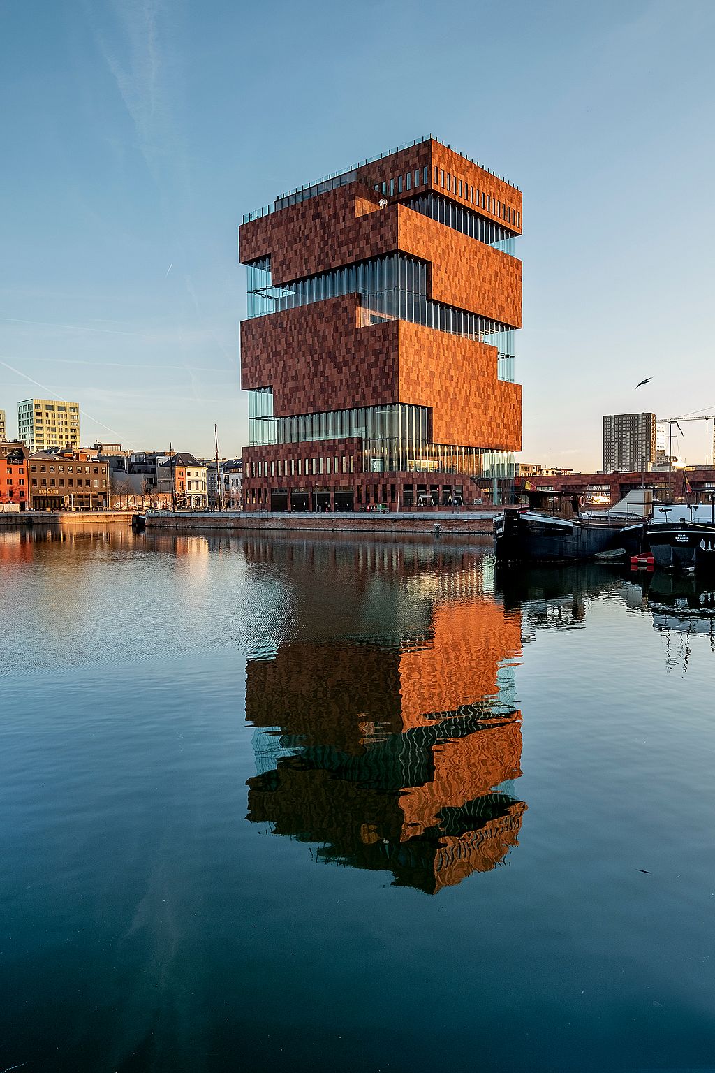 Außenansicht des MAS-Museums in Antwerpen, reflektiert im ruhigen Wasser der Docks unter klarem Abendhimmel.