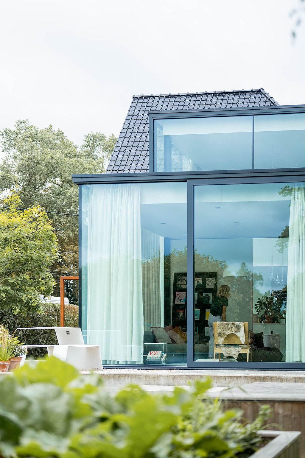 Vertical view of the glass rear façade of the house with a white Picnik table outside, seen across a lush garden with broad green leaves in the foreground.