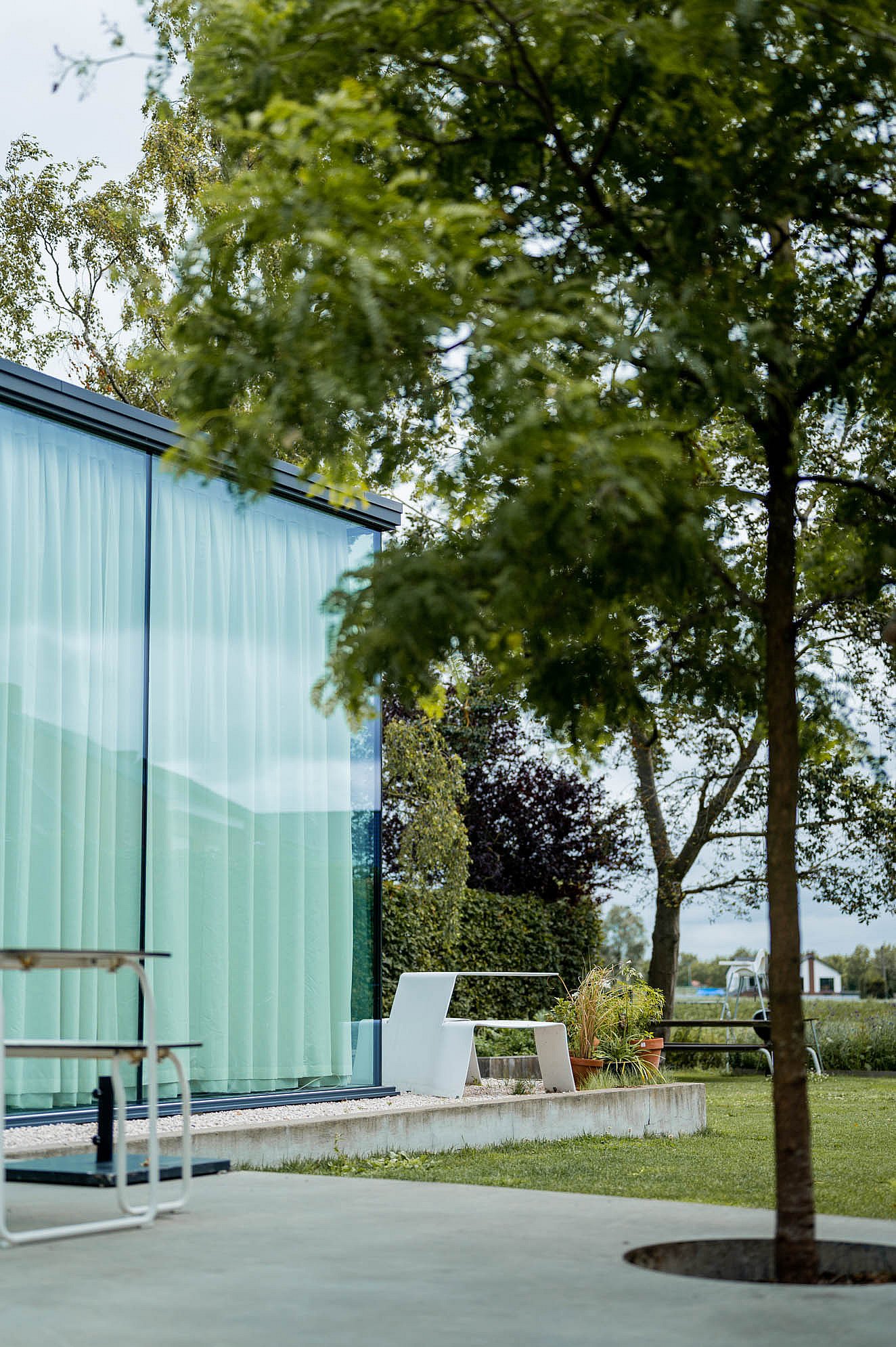 Seen through overhanging tree branches, a white Picnik table stands beside the glazed corner of the house overlooking the lawn and open landscape.