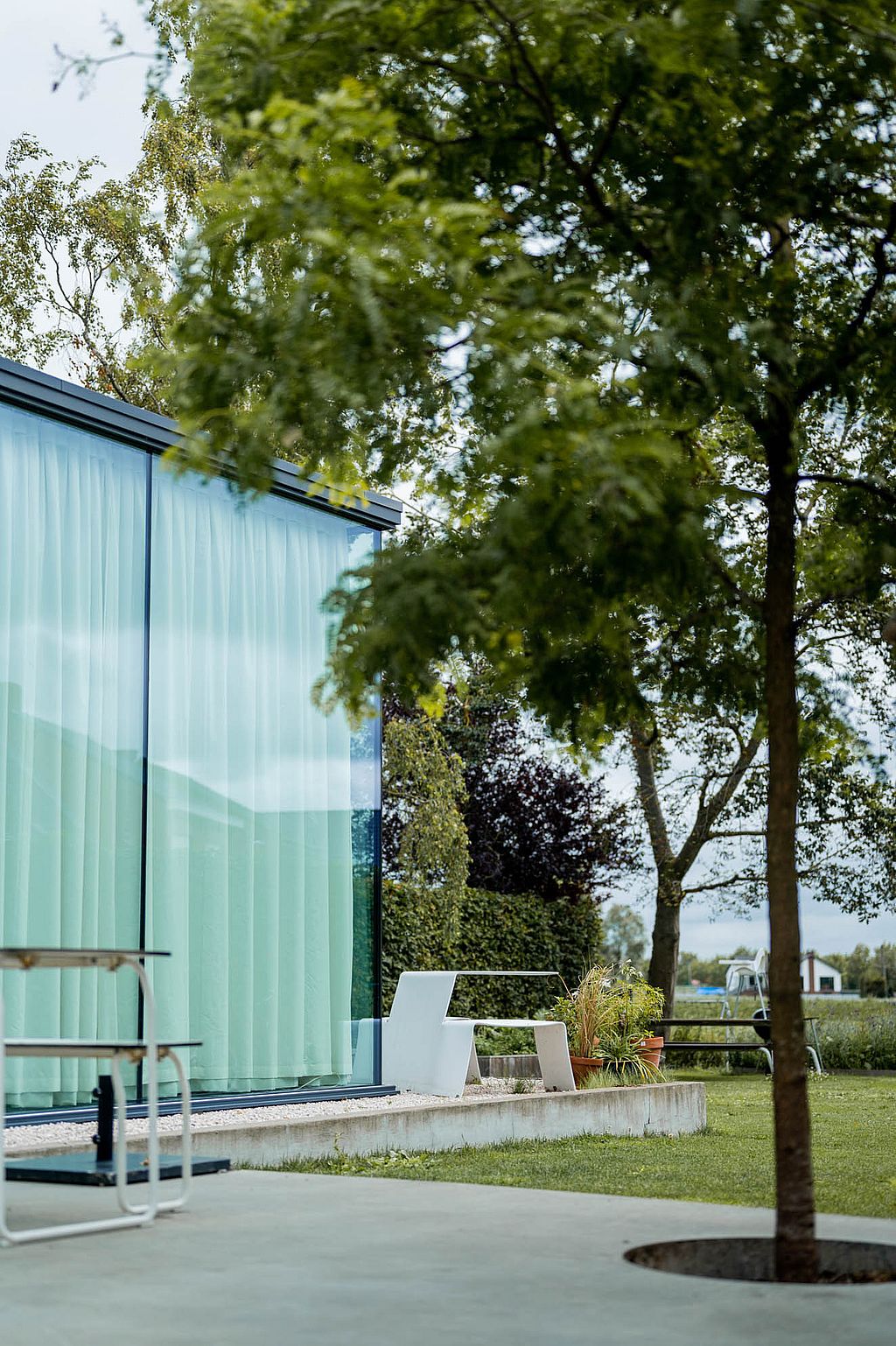 Seen through overhanging tree branches, a white Picnik table stands beside the glazed corner of the house overlooking the lawn and open landscape.