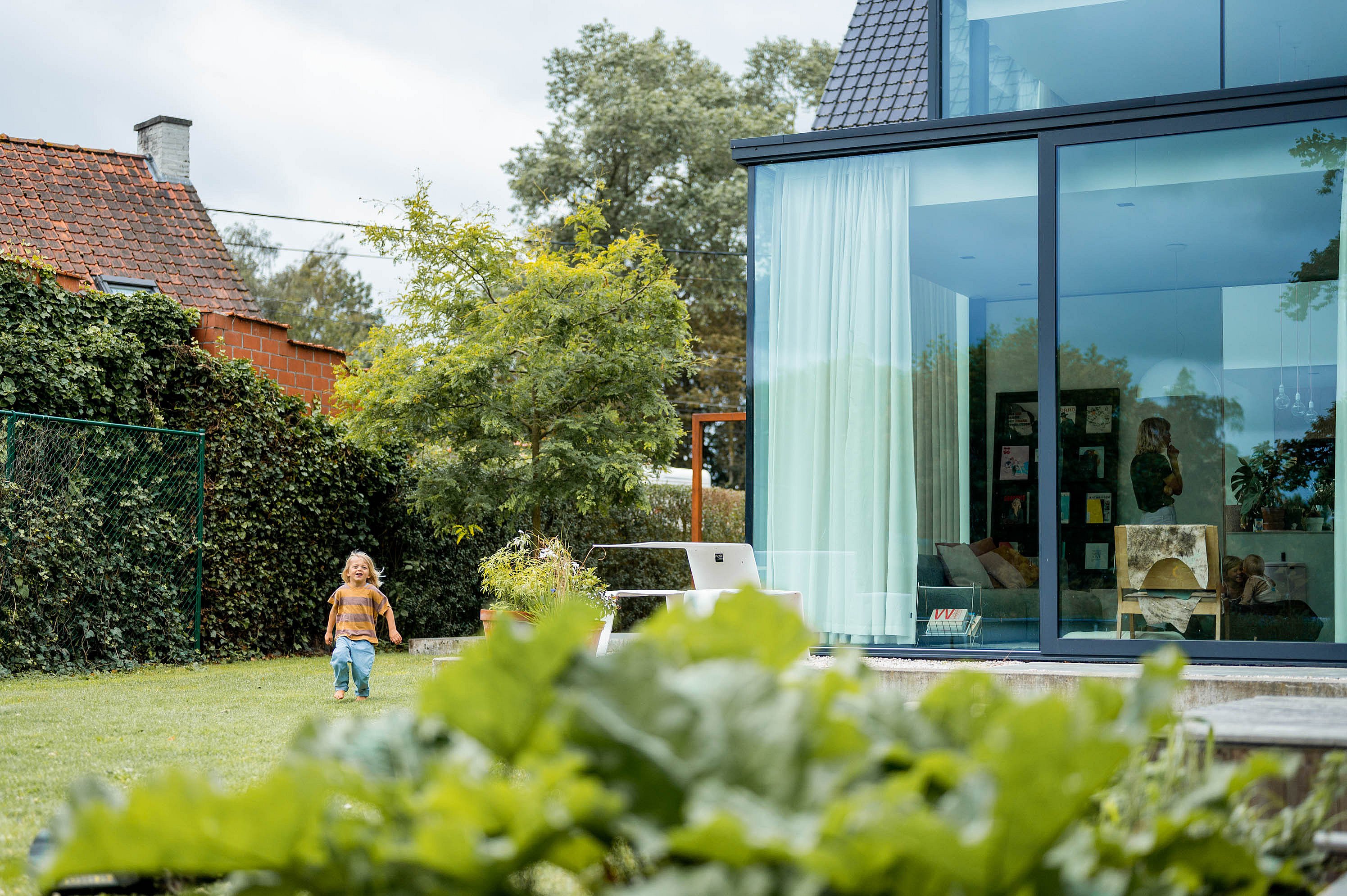 Wide garden view of a modern glass extension with a white Picnik table on the terrace, while a small child runs across the lawn in the foreground.
