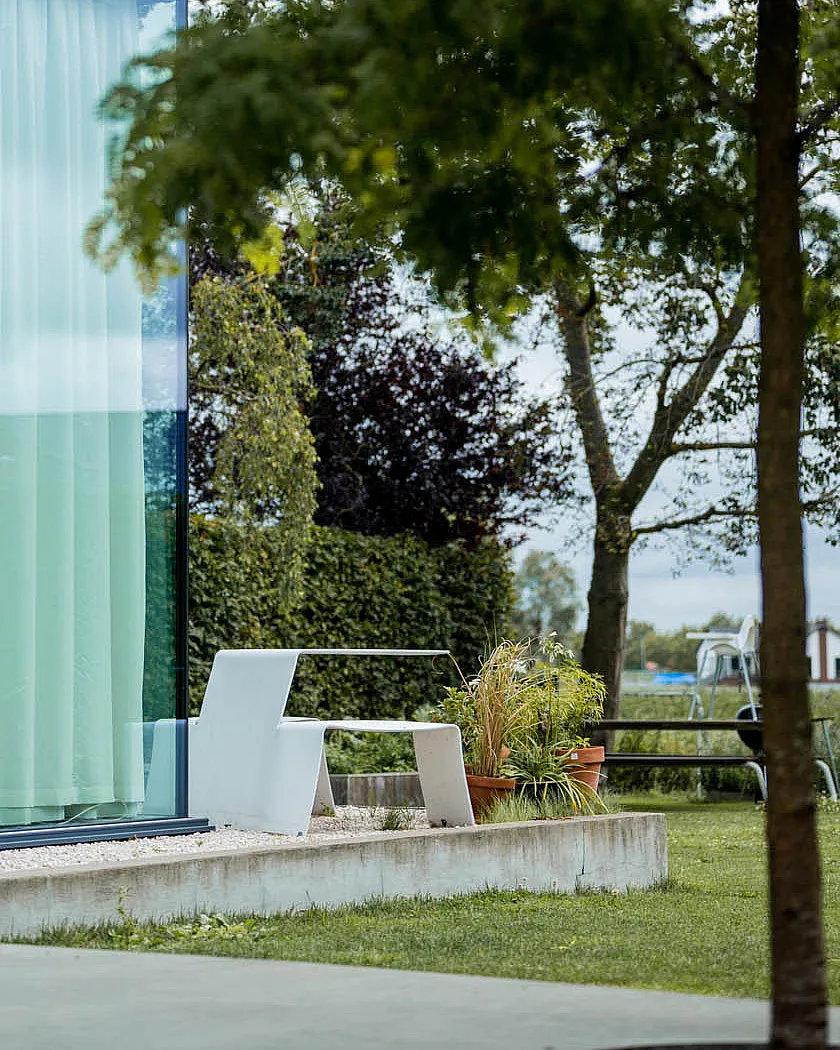 Een witte Picnik-tafel staat naast de glazen gevel van een modern huis, gelegen op een grindstrip met potplanten en een lommerrijke tuin daarachter.