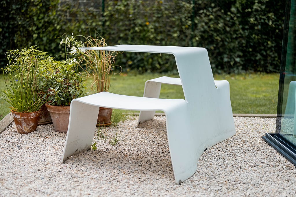 Close-up of a white Picnik table on pale gravel beside potted plants, highlighting its clean bent-aluminium form in the garden.