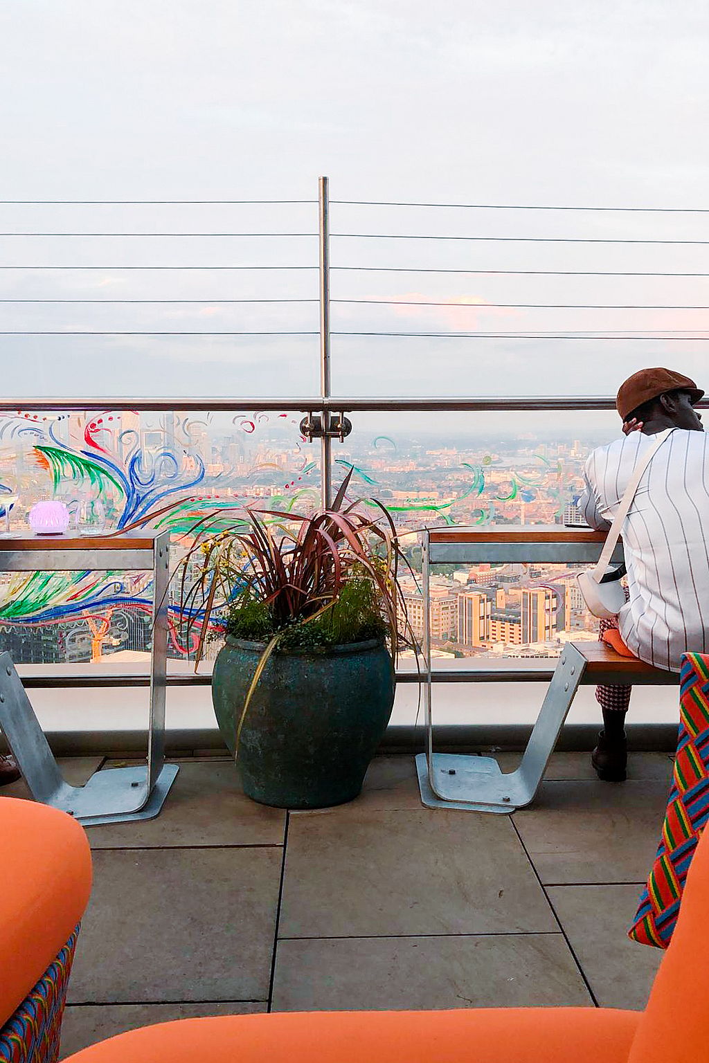 Un invité est assis sur un banc Hopper orangé au bord de la terrasse sur le toit, surplombant Londres à travers la rampe en verre.