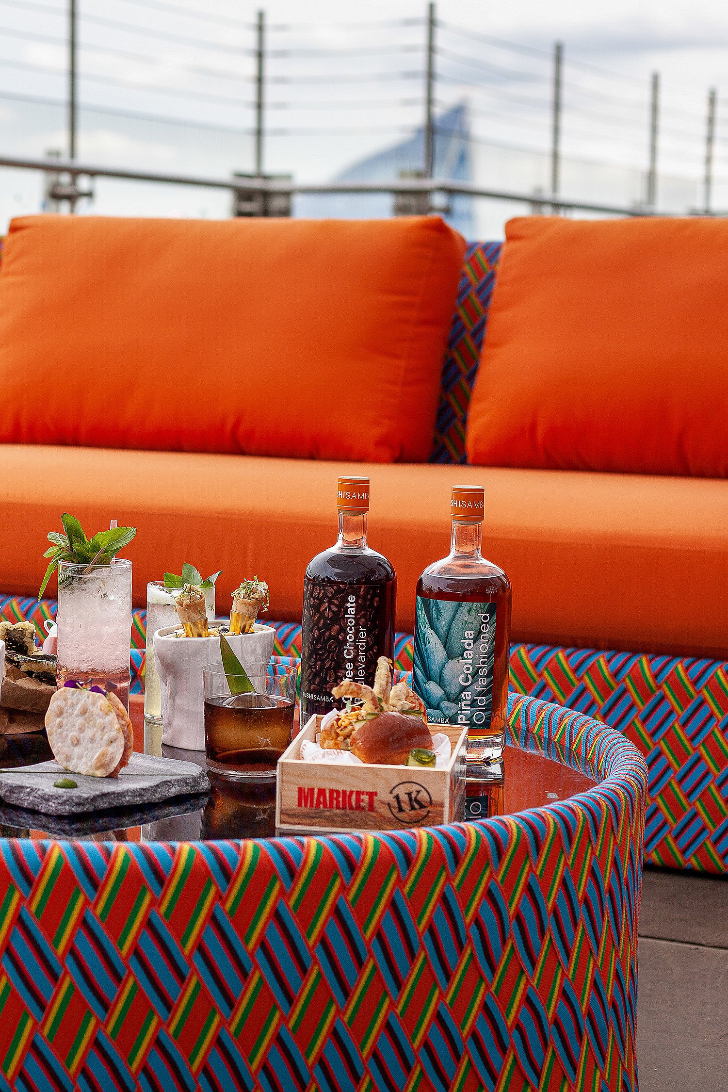 Gros plan de cocktails et petits plats sur une table ronde colorée devant des sièges lounge orange vif sur le toit-terrasse.