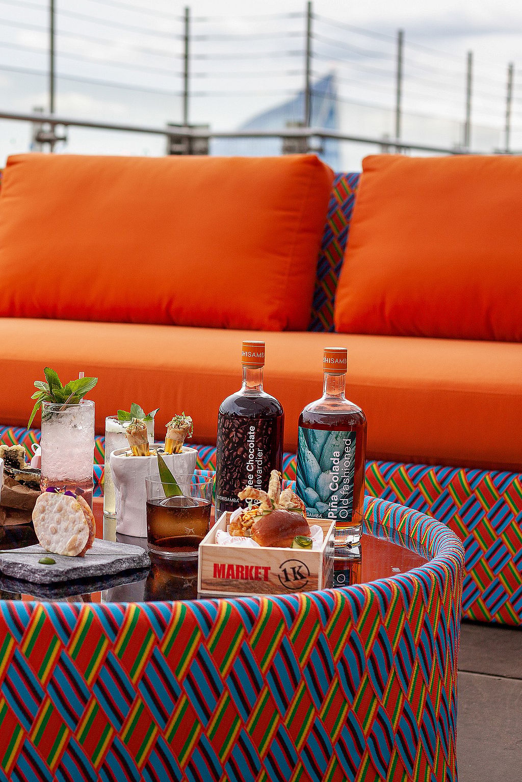 Gros plan de cocktails et petits plats sur une table ronde colorée devant des sièges lounge orange vif sur le toit-terrasse.