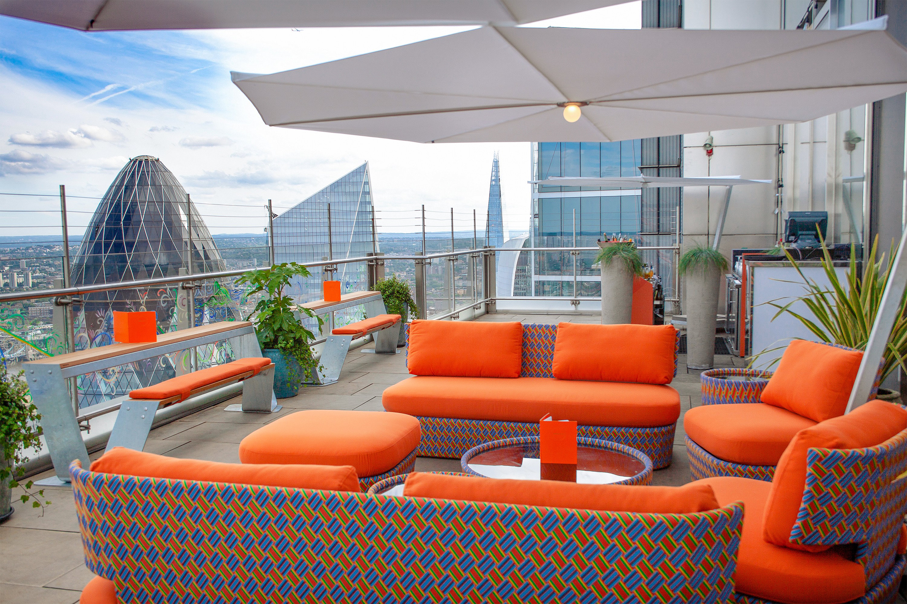 Salon coloré sur le toit avec des sièges incurvés orange vif, un grand parasol blanc et des bancs Hopper le long de la balustrade en verre, situé haut au-dessus de l’horizon de Londres.