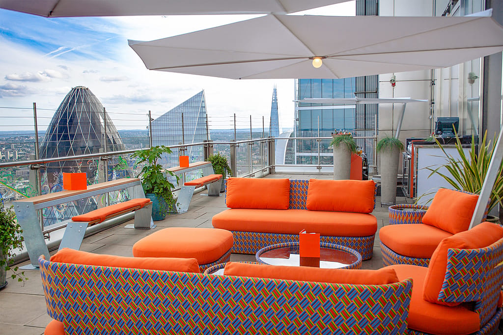Salon coloré sur le toit avec des sièges incurvés orange vif, un grand parasol blanc et des bancs Hopper le long de la balustrade en verre, situé haut au-dessus de l’horizon de Londres.