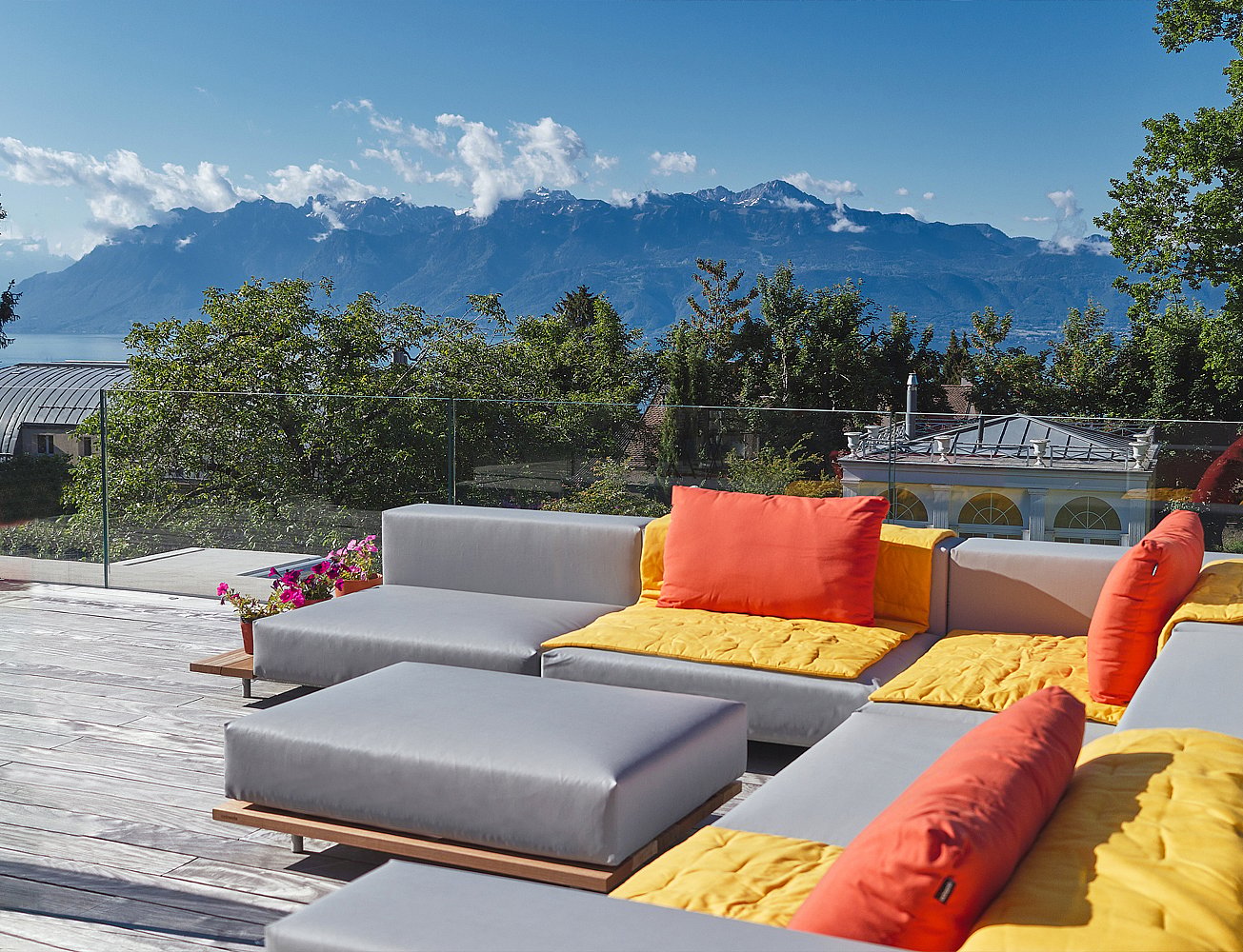 Ein graues modulares Walrus-Außensofa mit gelben Sitzpolstern und orangefarbenen Rückenpolstern steht auf einer Holzterrasse mit Blick auf den Lémansee und die Schweizer Berge.