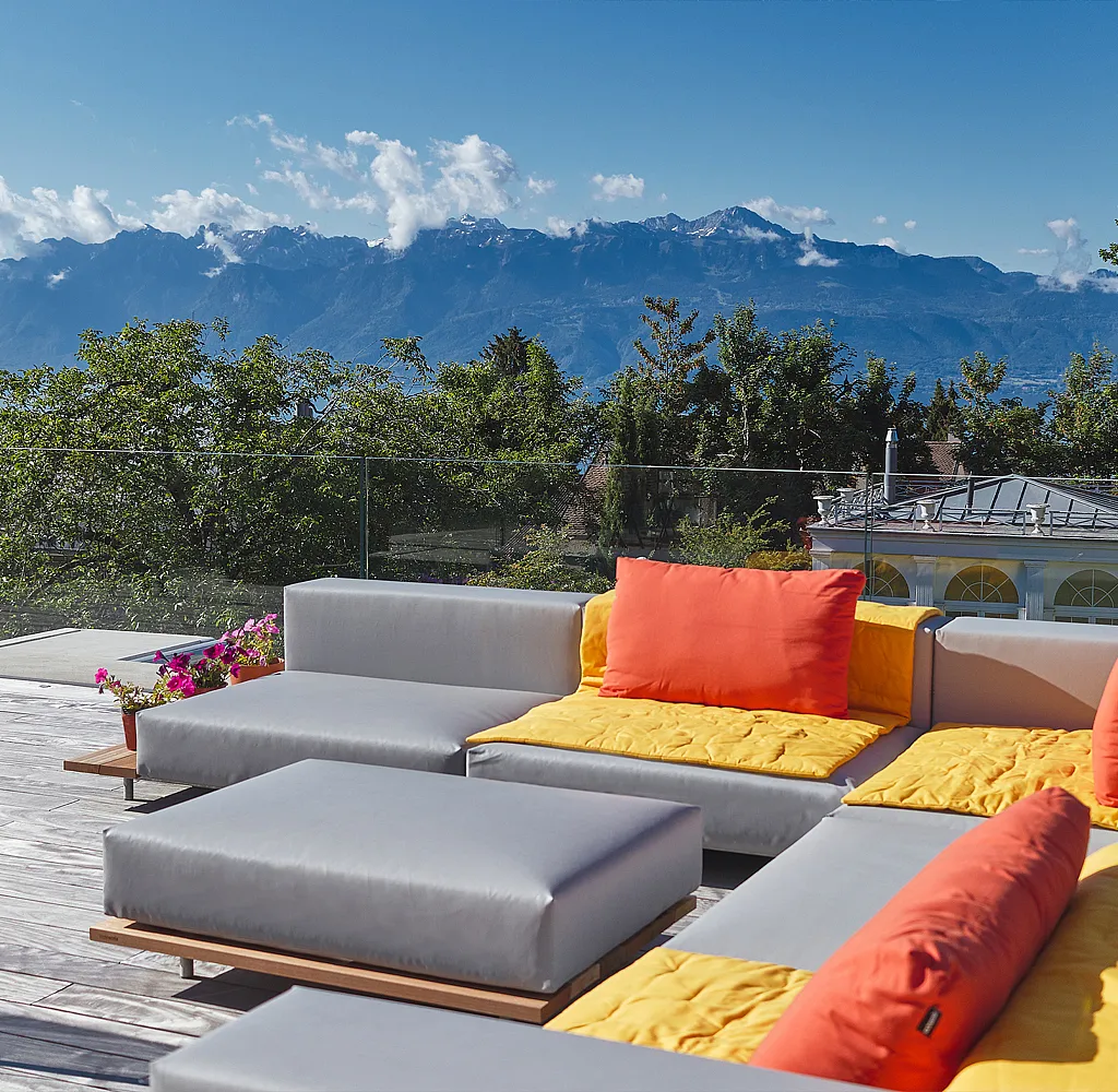 Ein graues modulares Walrus-Außensofa mit gelben Sitzpolstern und orangefarbenen Rückenpolstern steht auf einer Holzterrasse mit Blick auf den Lémansee und die Schweizer Berge.
