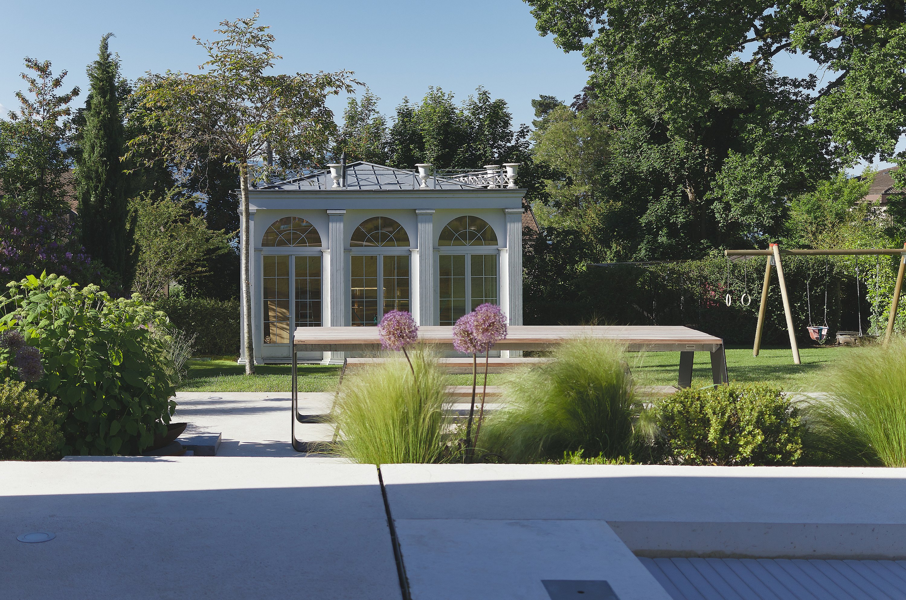 Ein langer Hopper-Picknicktisch steht in einem gepflegten Garten mit Ziergräsern, Bäumen und einem kleinen klassischen Gartenpavillon im Hintergrund.