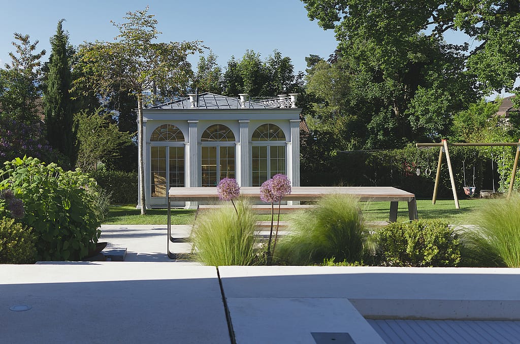 Ein langer Hopper-Picknicktisch steht in einem gepflegten Garten mit Ziergräsern, Bäumen und einem kleinen klassischen Gartenpavillon im Hintergrund.