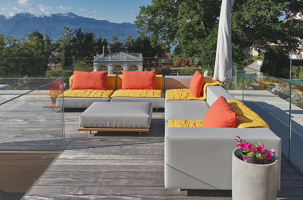 Ein L-förmiges Walrus-Außensofa mit gelben Sitzpolstern, orangefarbenen Kissen und einem passenden Ottomanen steht auf einer Terrasse mit Glasgeländer und Bergblick.