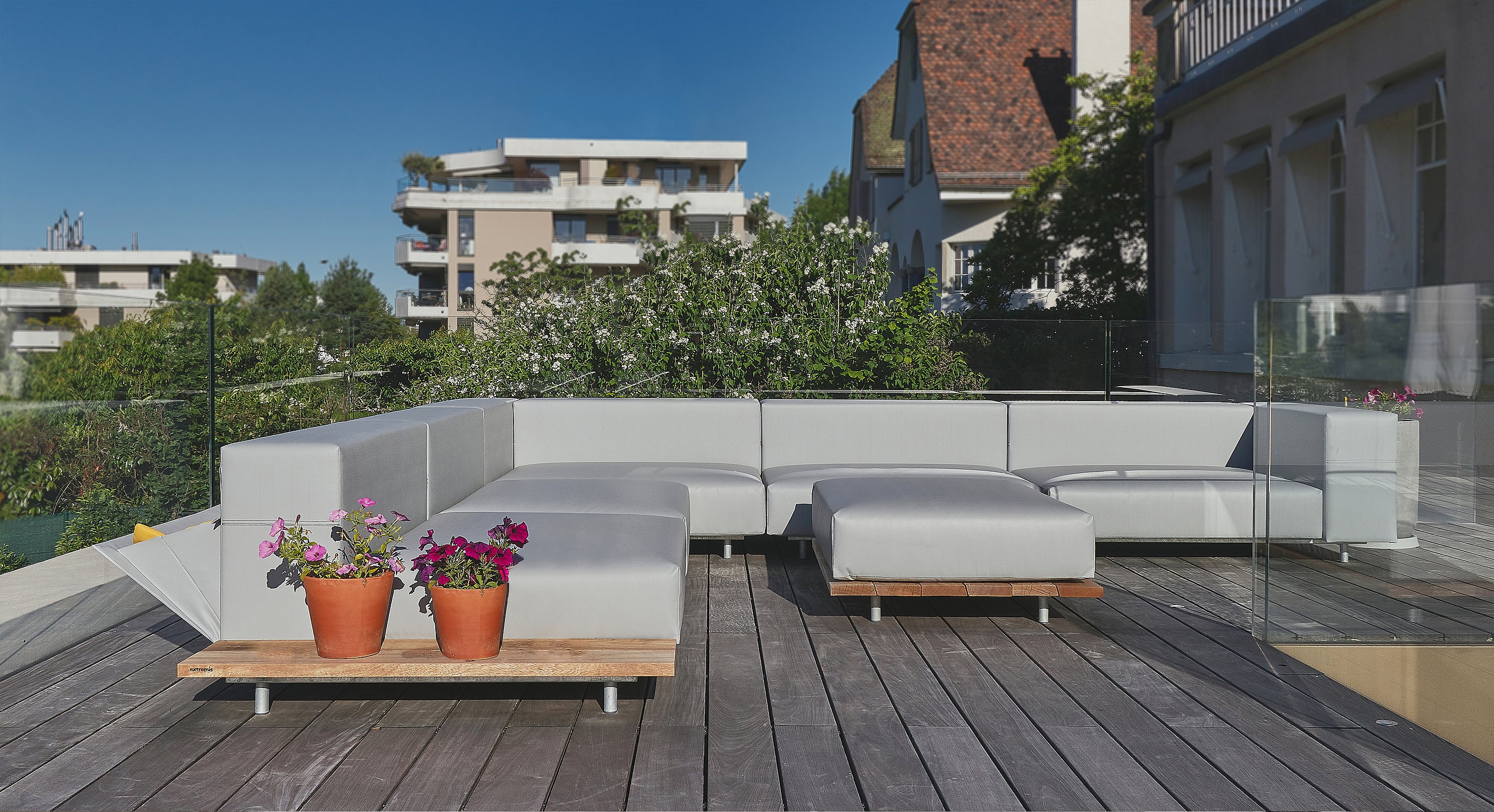 Ein leeres modulares Walrus-Sofa mit Ottomanen steht auf einer geräumigen Holzterrasse mit Glasbalustraden, Topfblumen und Blick auf den grünen Garten.