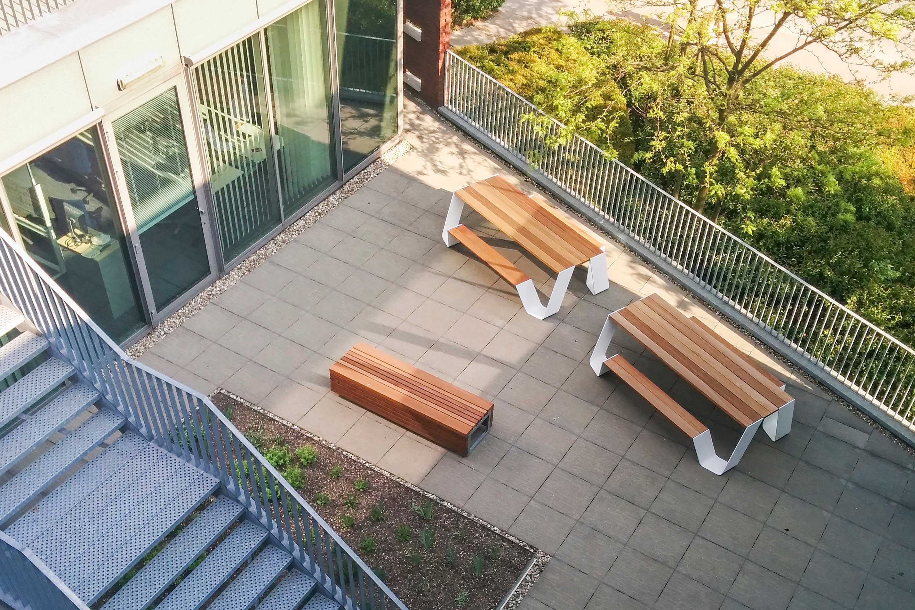Vue aérienne d’un bureau sur le toit depuis la terrasse telenet avec deux tables de pique-nique Hopper blanches et un banc assorti à côté du bâtiment Telenet, bordé par des balustrades et des arbres feuillus.