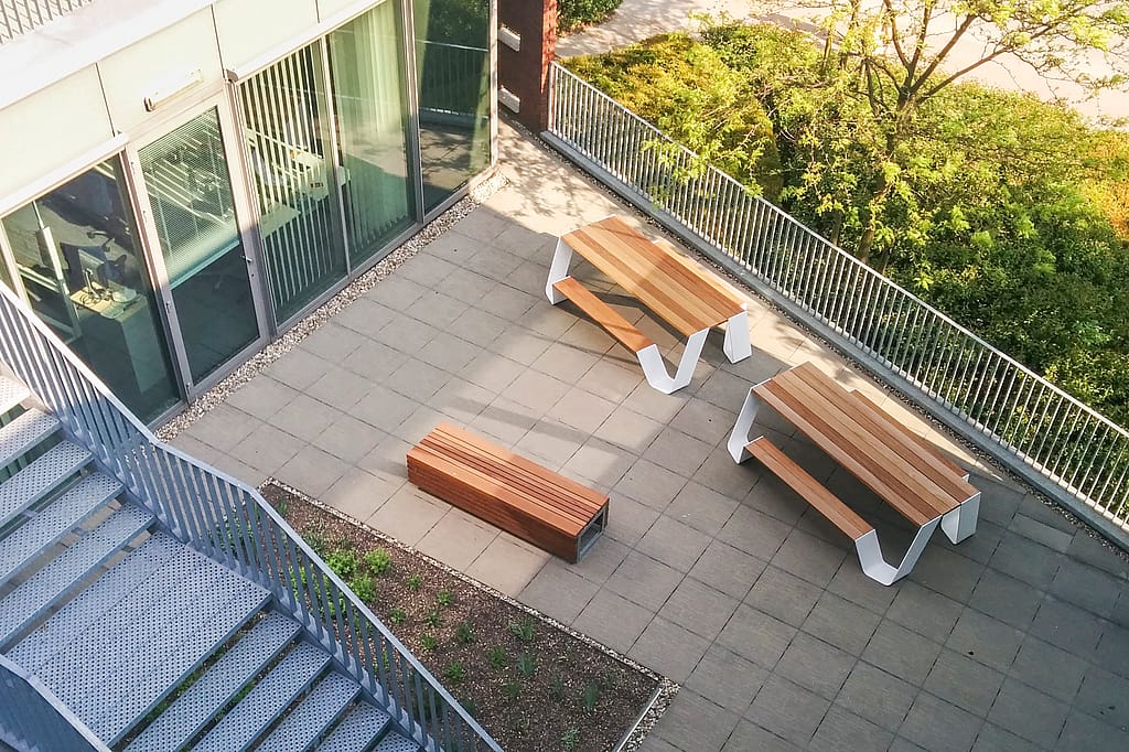 Draufsicht auf eine Büroterrasse auf dem Dach mit zwei weißen Hopper-Picknicktischen und einer passenden Bank neben dem Telenet-Gebäude, gesäumt von Geländern und grünen Bäumen.