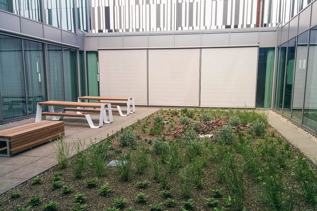 Ein geschlossener Bürohof mit zwei weißen Hopper-Picknicktischen neben einem bepflanzten zentralen Bett, umgeben von hohen Glaswänden und einer leichten Metallverkleidung.