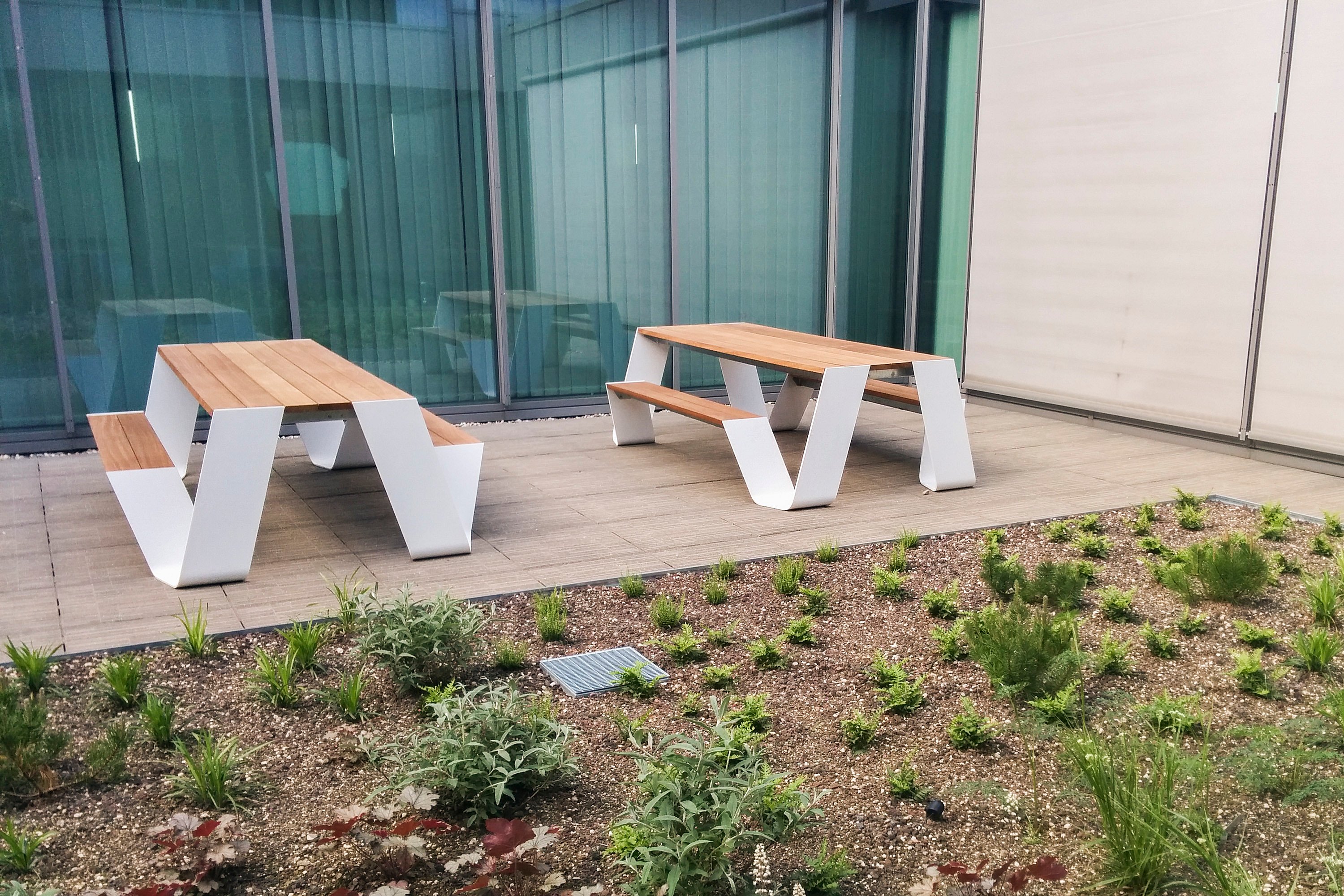 Zwei weiße Hopper-Picknicktische stehen auf einer Holzterrasse in einem kompakten Terrassengarten, neben raumhohen Fenstern und niedrigem Grün.