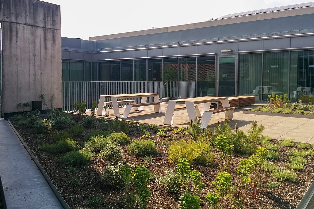 Zwei weiße Hopper-Picknicktische stehen auf einer sonnenbeschienenen Terrasse vor den Telenet-Büros, eingerahmt von niedrigen Beeten, Geländern und einer Glasfassade.