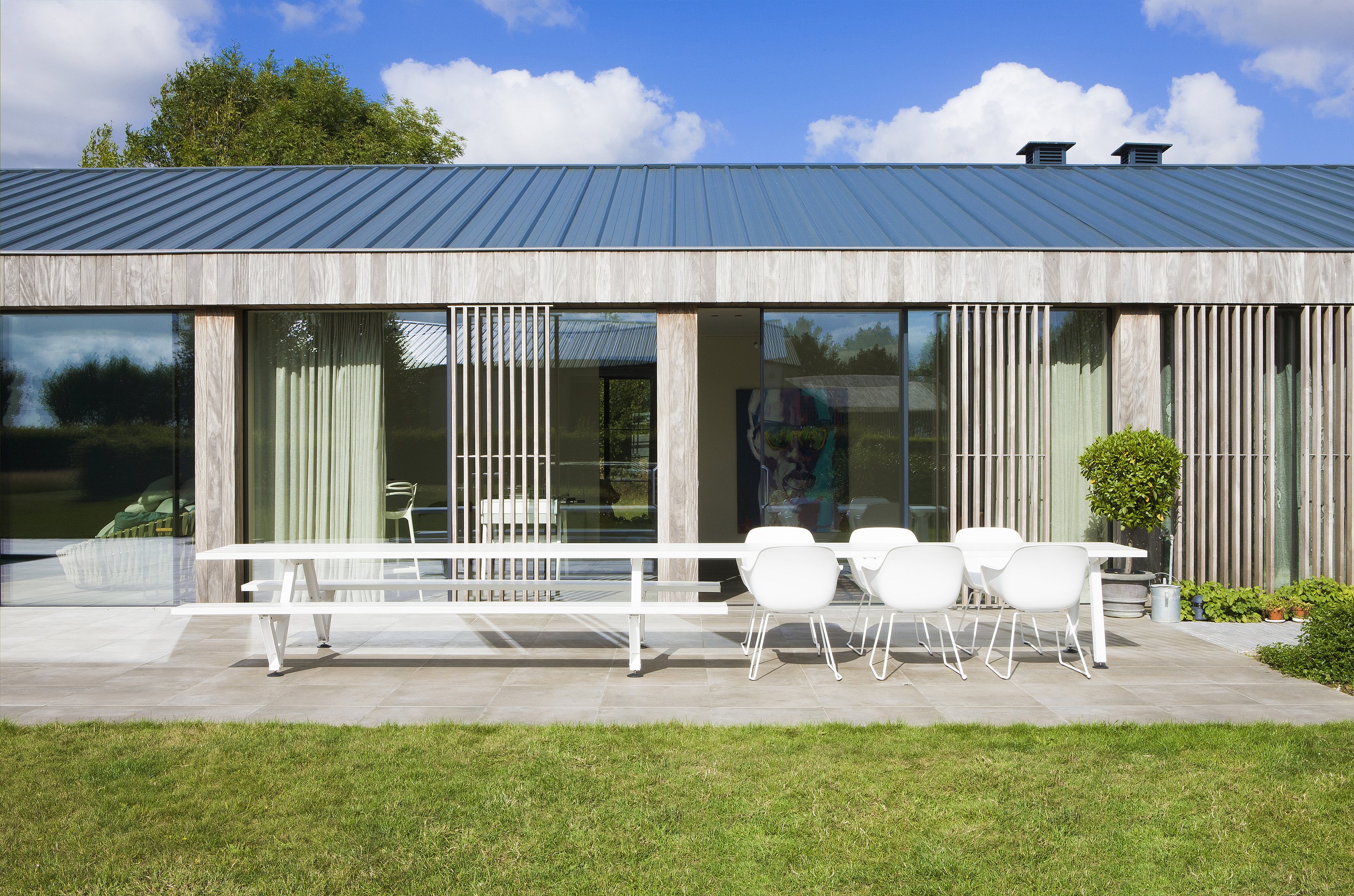 Vue de face d’une longue marina blanche combinée avec des bancs et des chaises sur la terrasse devant la façade vitrée de la maison.