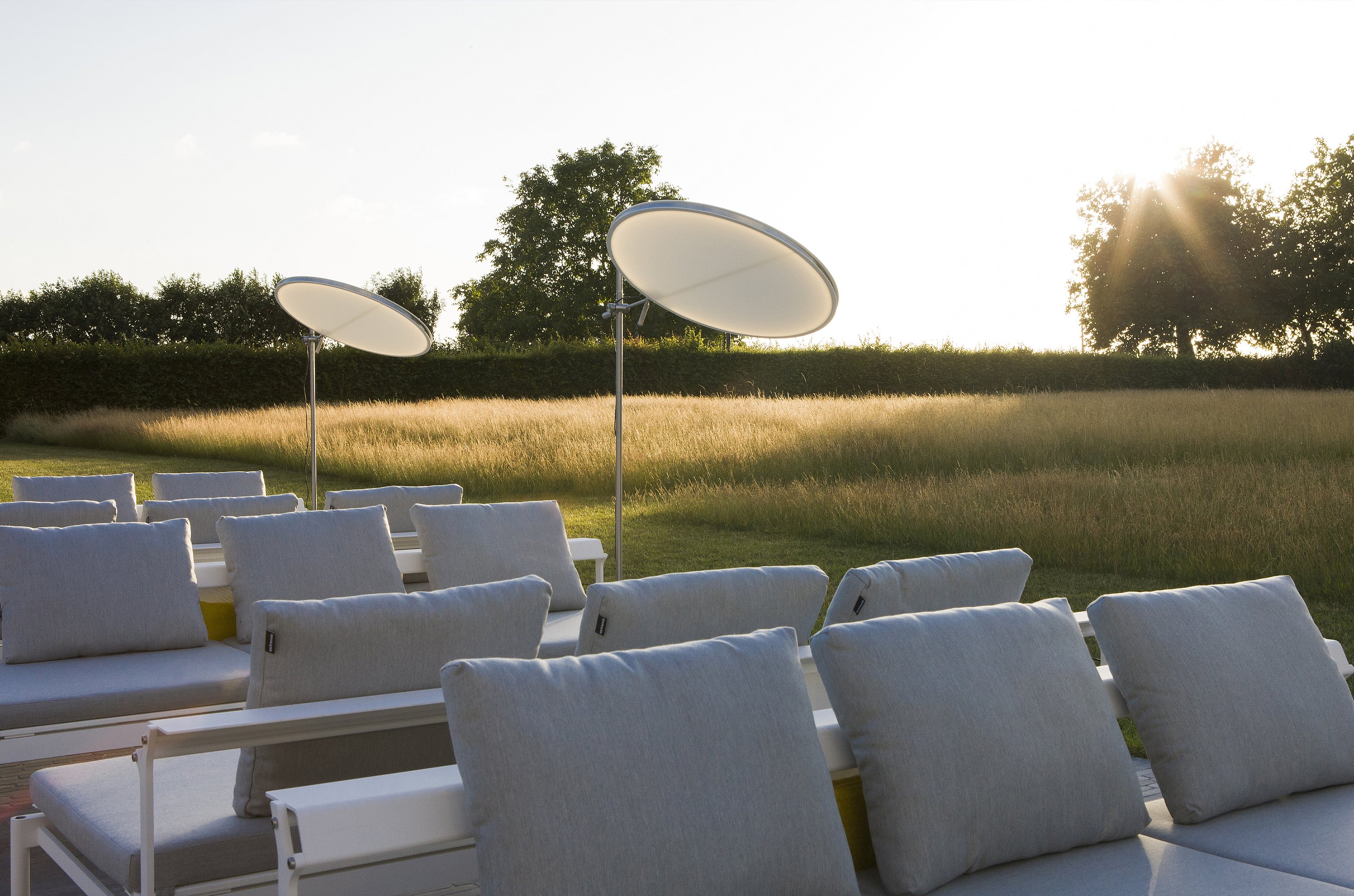 Gros plan de coussins arrière pâles sur des chaises longues blanches Sol+Luna, avec le soleil du soir bas brillant à travers la prairie au-delà.