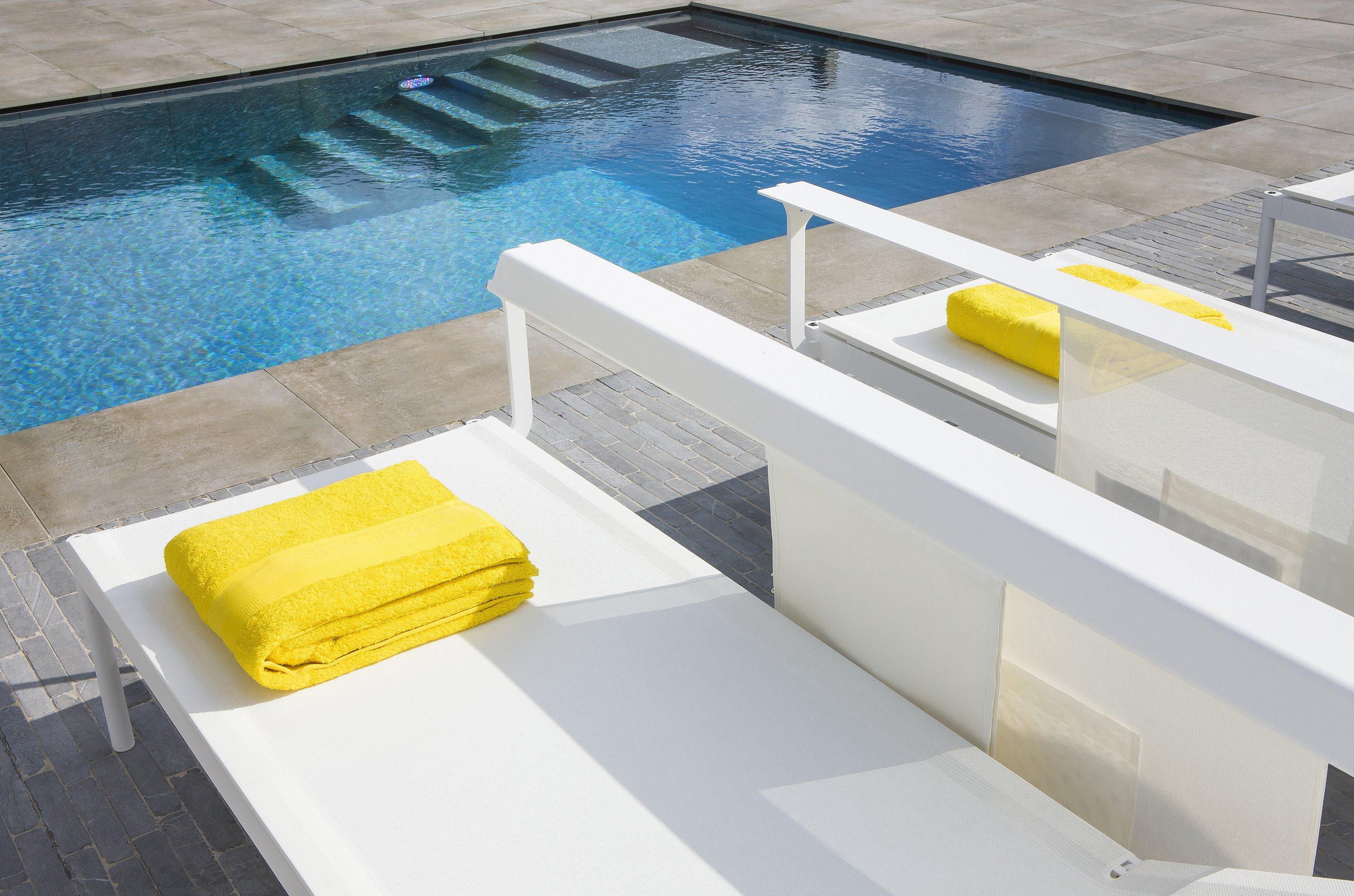 Gros plan de deux transats blancs Sol+Luna au bord de la piscine, chacun surmonté d’une serviette pliée jaune vif.