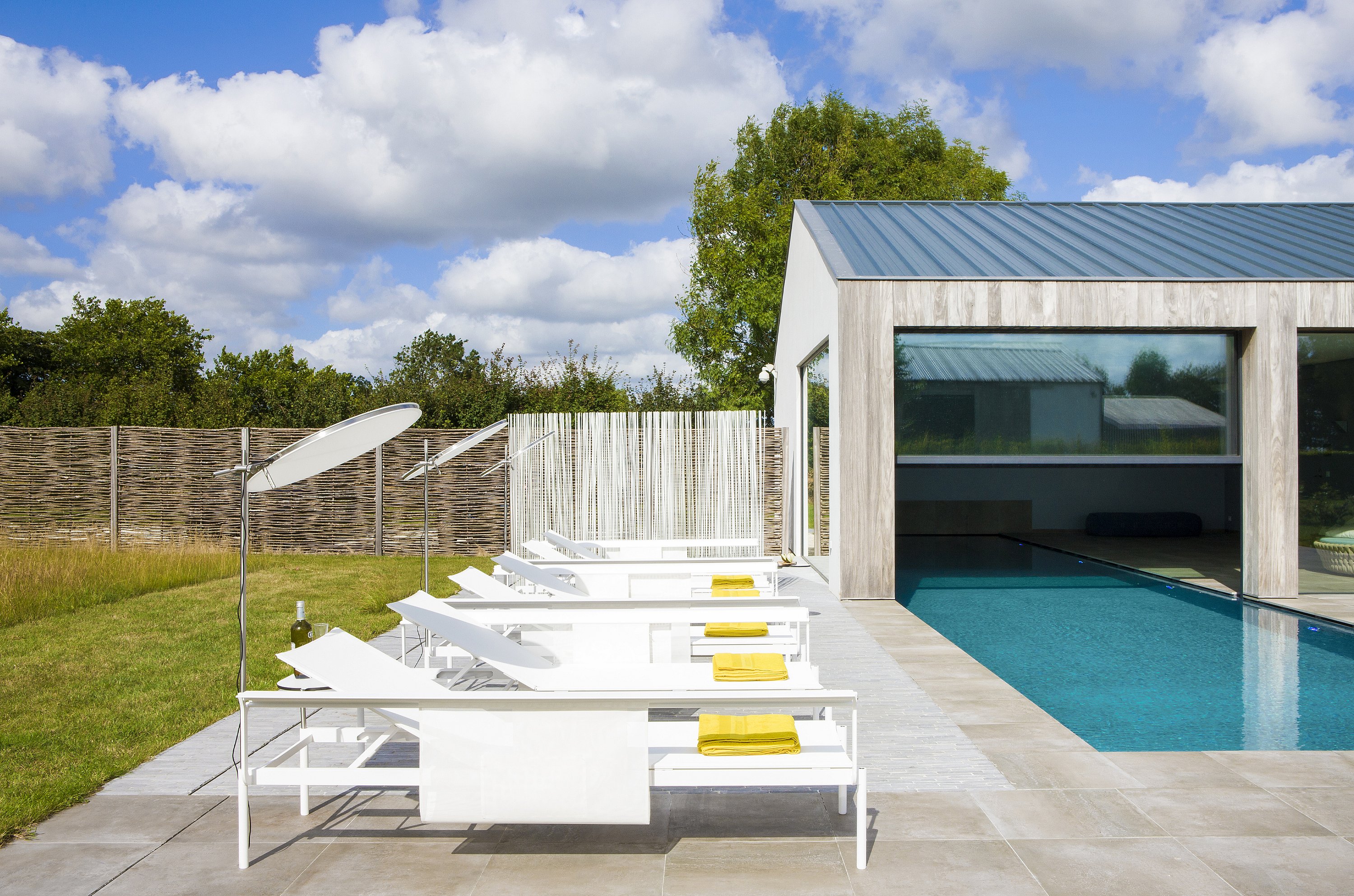 Vue latérale de plusieurs chaises longues blanches Sol+Luna disposées à côté de la piscine de la maison de vacances en bois, avec des serviettes jaunes ajoutant du contraste.