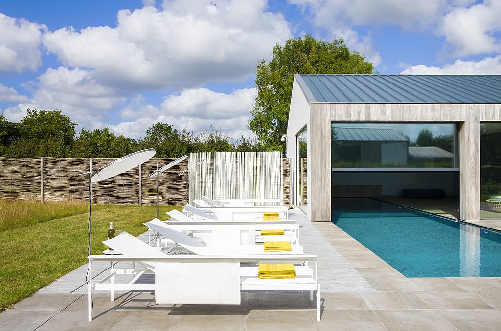 Vue latérale de plusieurs chaises longues blanches Sol+Luna disposées à côté de la piscine de la maison de vacances en bois, avec des serviettes jaunes ajoutant du contraste.
