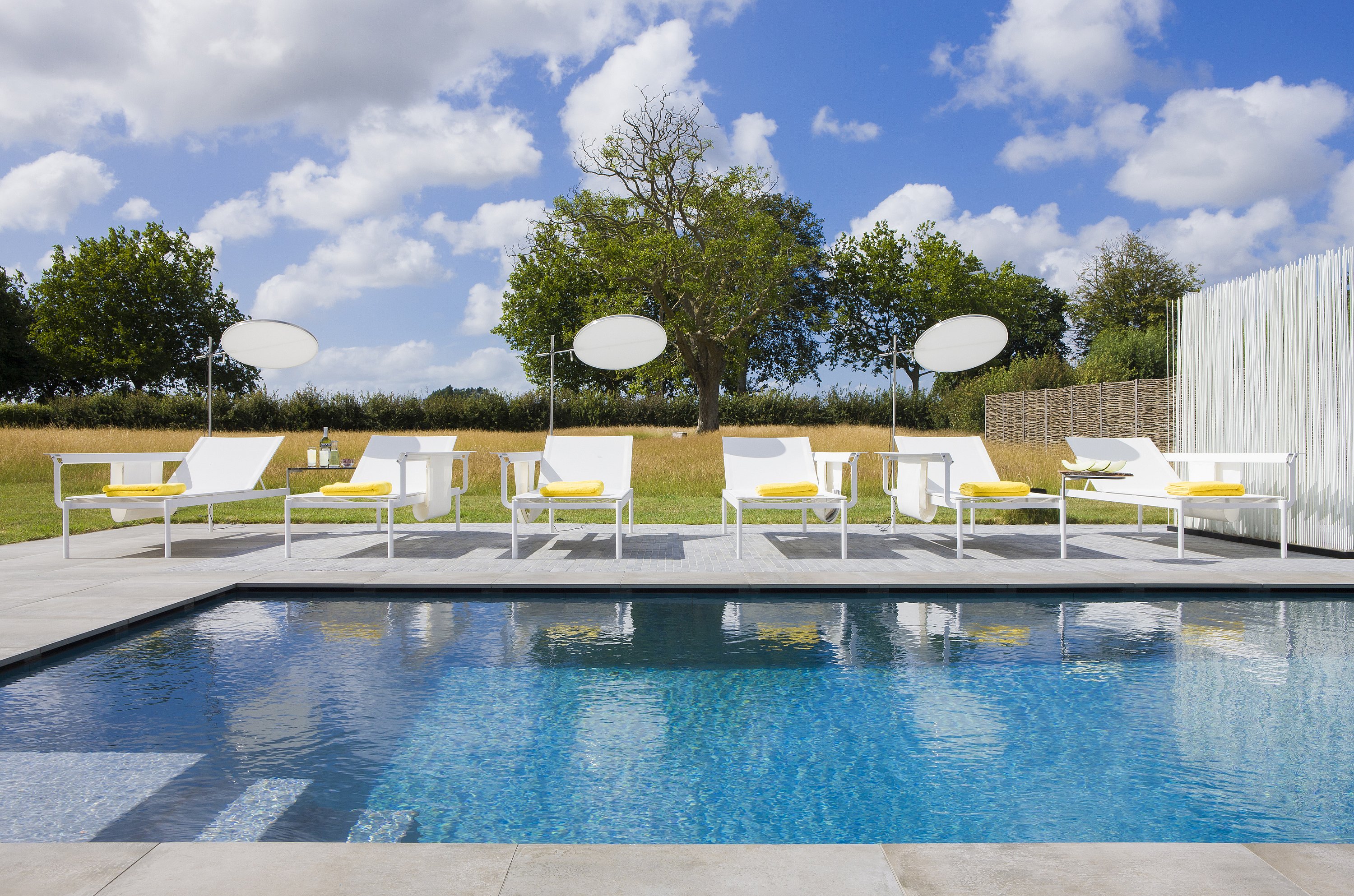 Eine Reihe weißer Sol+Luna-Liegen mit leuchtend gelben Handtüchern säumt den Rand eines Swimmingpools, eingebettet in eine Graslandschaft und einen blauen Himmel mit vereinzelten Wolken.