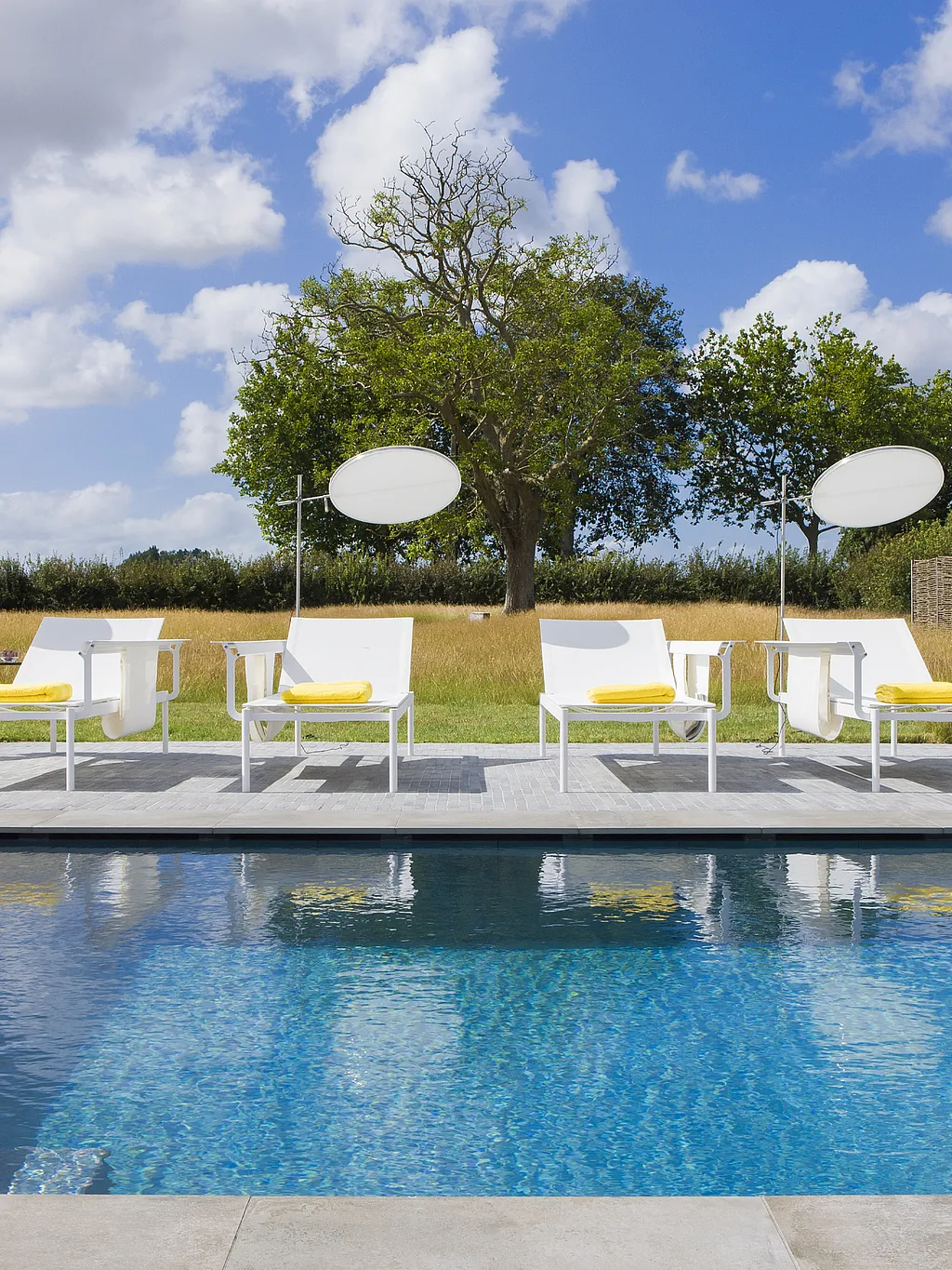 Eine Reihe weißer Sol+Luna-Liegen mit leuchtend gelben Handtüchern säumt den Rand eines Swimmingpools, eingebettet in eine Graslandschaft und einen blauen Himmel mit vereinzelten Wolken.
