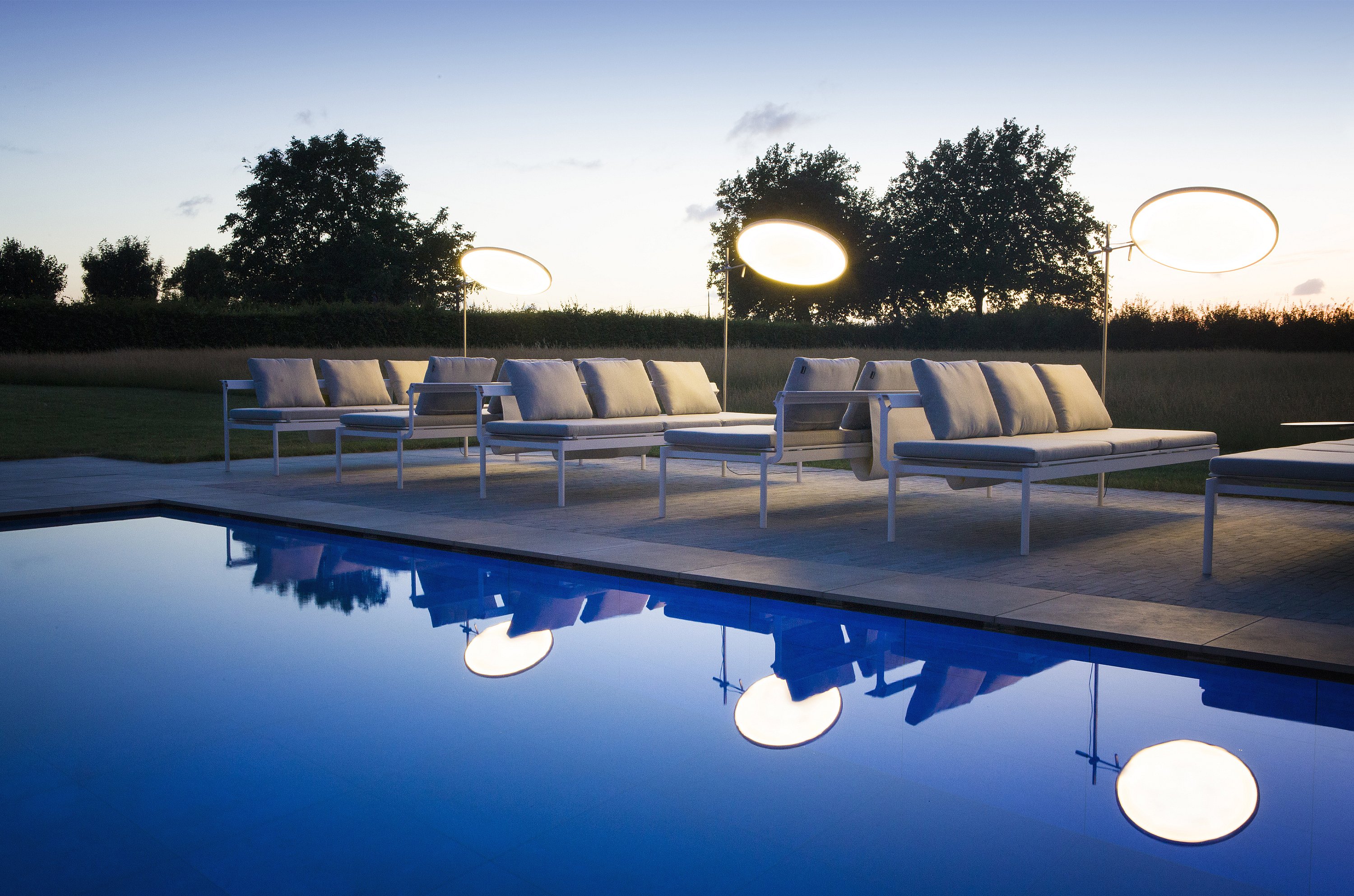 Les chaises longues blanches Sol+Luna sont assises à côté de la piscine au crépuscule, reflétées dans l’eau calme tandis que les canopées d’ombre rondes brillent contre le ciel du soir.