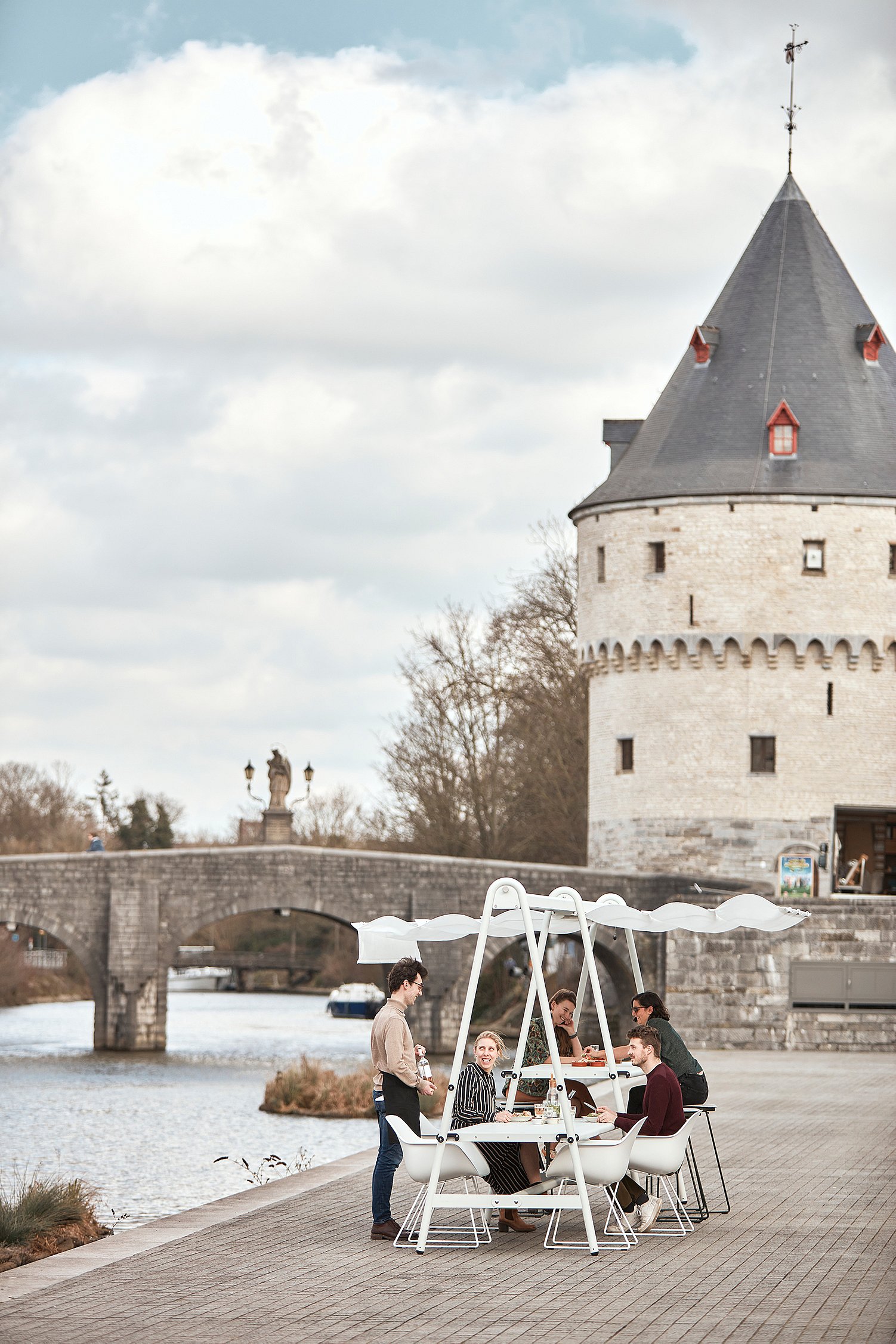 Fünf Personen sitzen und stehen um einen verbundenen weißen AMAi-Tisch am Fluss, hinter dem sich die Steinbrücke und einer der Broel Towers erheben.