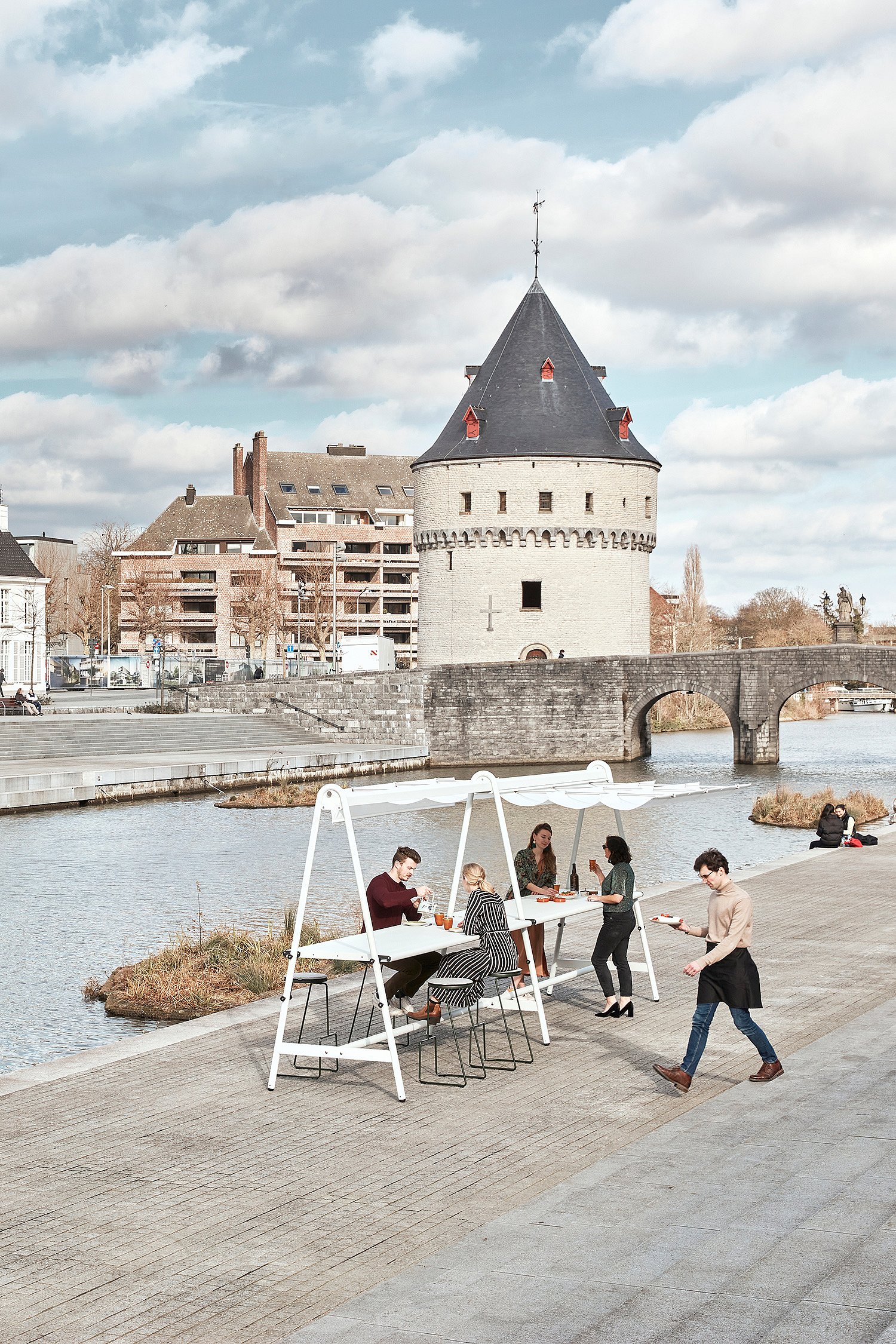 Eine Gruppe versammelt sich um ein weißes AMAi-Setup an einer Uferpromenade, mit weißen Kapitänsstühlen, schwarzen Tiki-Hockern und den Broel Towers von Kortrijk im Hintergrund.
