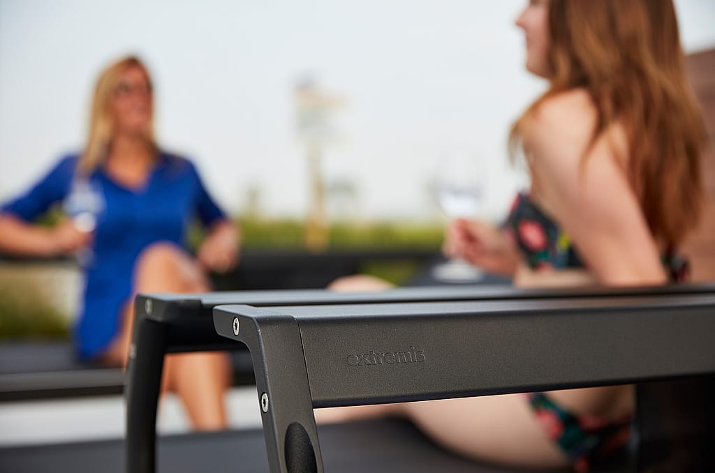 Gros plan sur la structure métallique noire d'un transat Sol+Luna. Deux femmes, un verre à la main, apparaissent légèrement floues en arrière-plan.