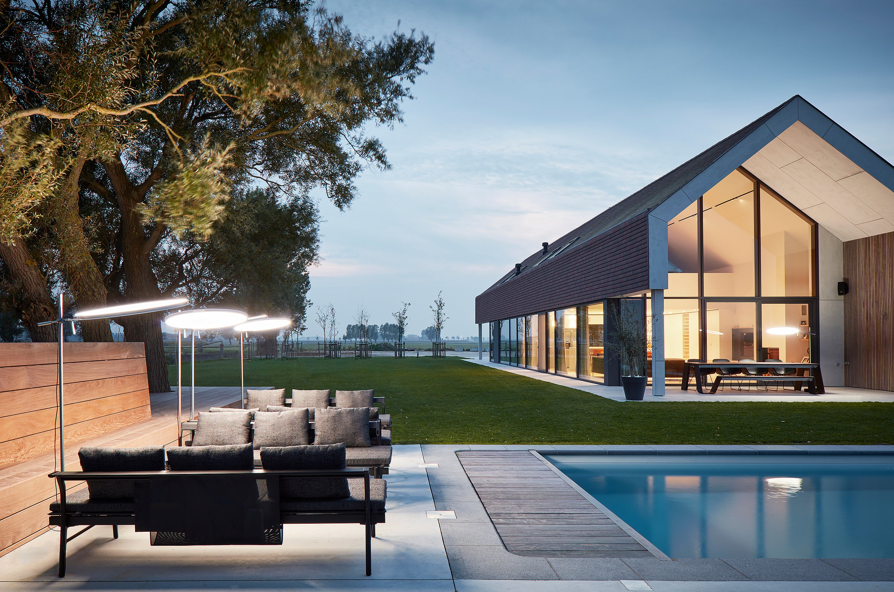 Au crépuscule, des transats Sol+Luna anthracite, coiffés de parasols ronds lumineux, bordent une piscine devant une maison moderne à pignon. Une table Hopper est visible sur la terrasse.