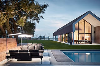 Au crépuscule, des transats Sol+Luna anthracite, coiffés de parasols ronds lumineux, bordent une piscine devant une maison moderne à pignon. Une table Hopper est visible sur la terrasse.