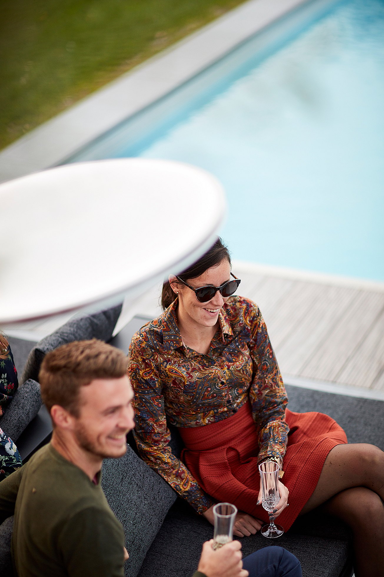 Vue aérienne d'amis se relaxant sur des chaises longues Sol+Luna au bord de la piscine, une coupe de champagne à la main, sous un parasol rond blanc.