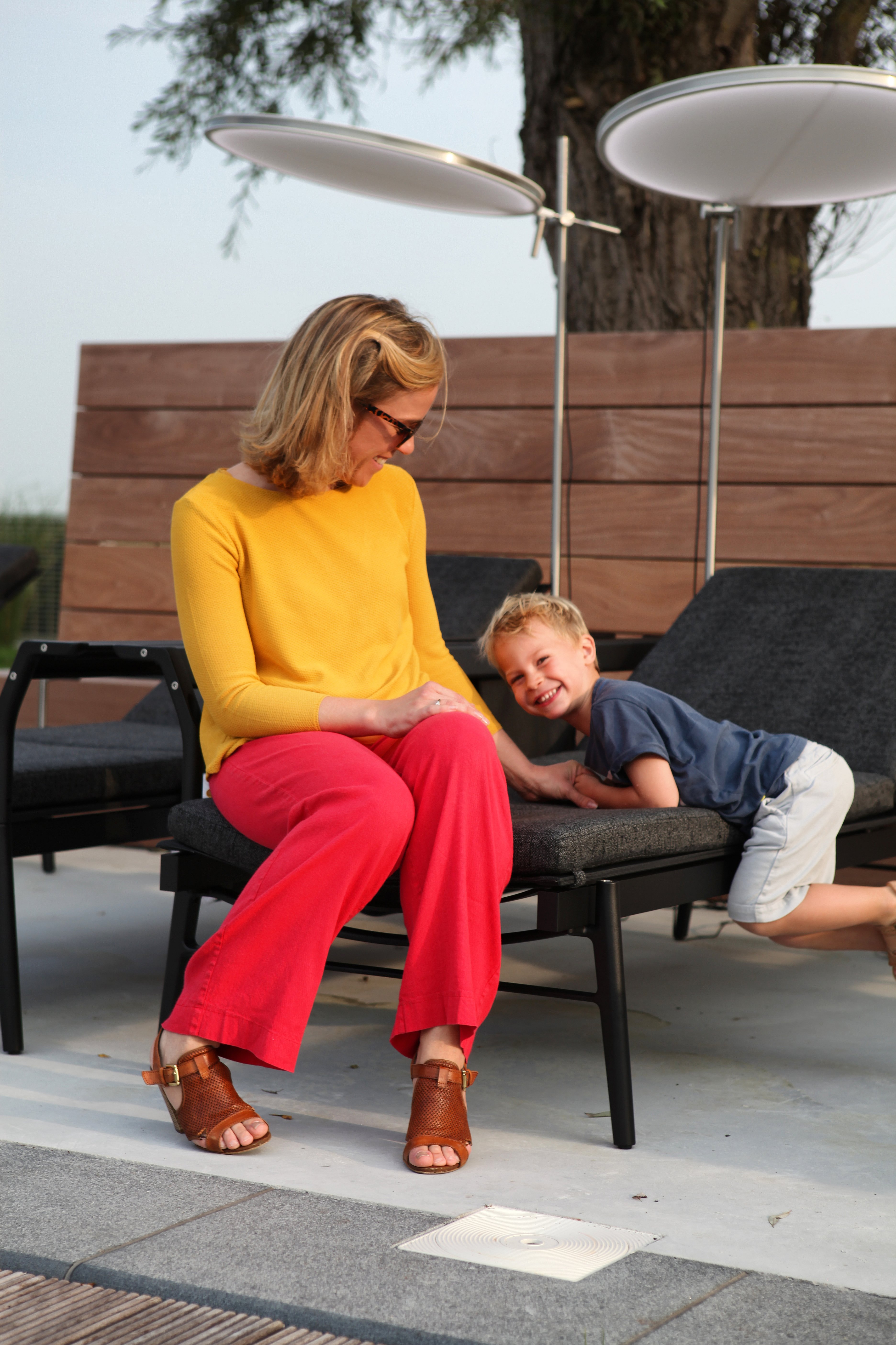 Une femme vêtue de couleurs vives est assise avec un enfant souriant sur un transat Sol+Luna noir, derrière lesquels se trouvent des parasols ronds illuminés.