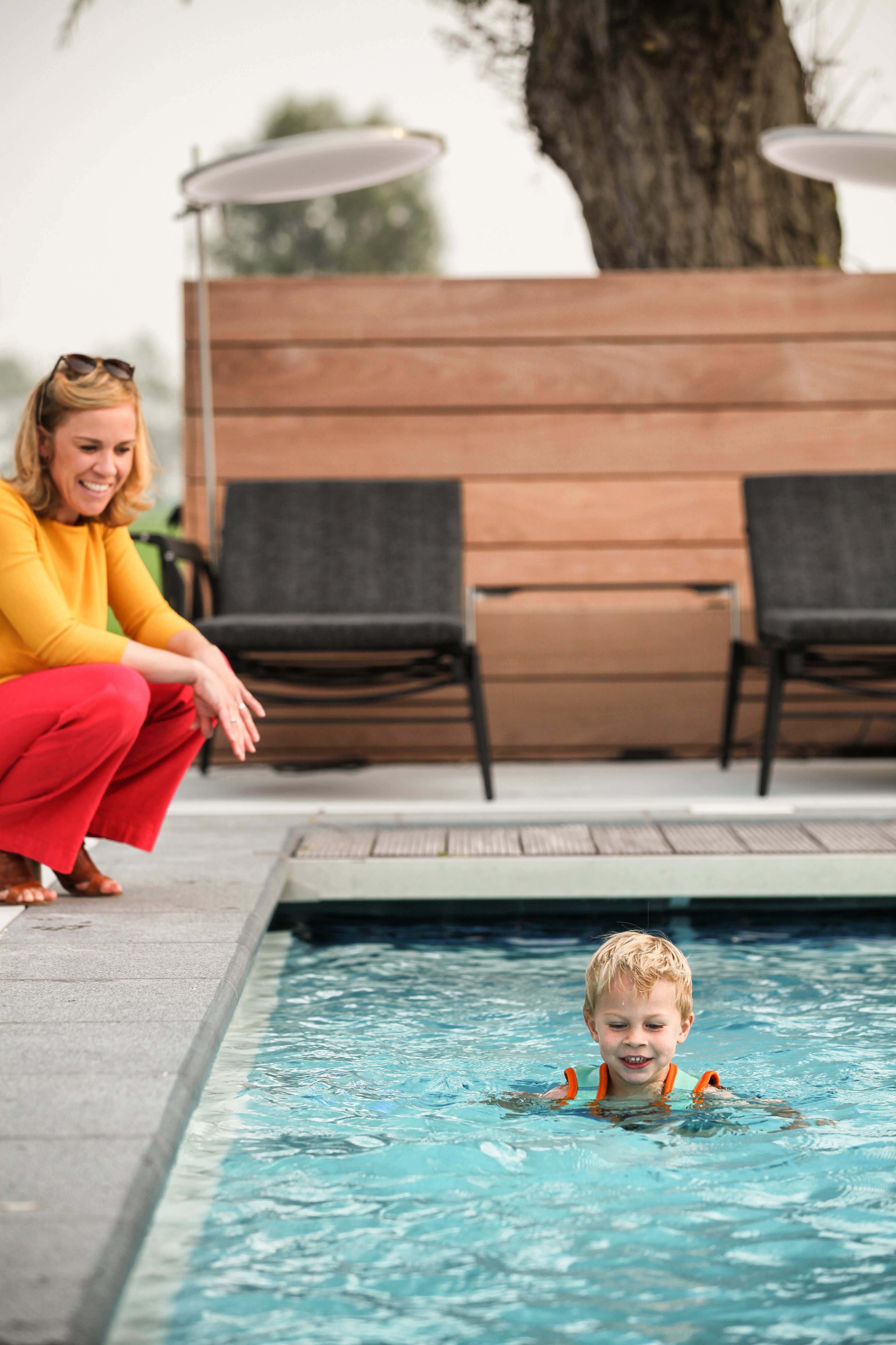   Un jeune garçon nage dans la piscine tandis qu'une femme l'observe depuis le bord. Derrière eux, on aperçoit des sièges Sol+Luna et des parasols ronds.