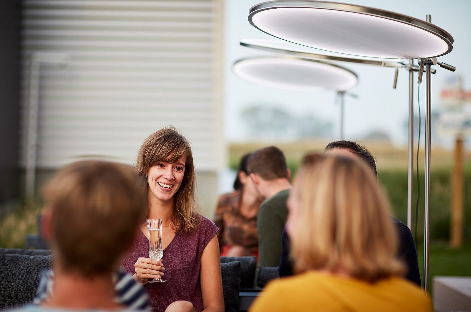 Des amis sont installés ensemble sur des chaises longues d'extérieur, sous des parasols ronds illuminés, et profitent d'un verre à la tombée du jour.