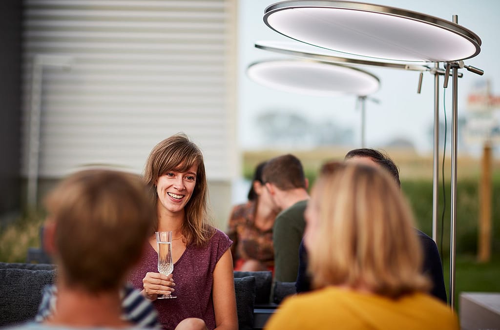 Des amis sont installés ensemble sur des chaises longues d'extérieur, sous des parasols ronds illuminés, et profitent d'un verre à la tombée du jour.