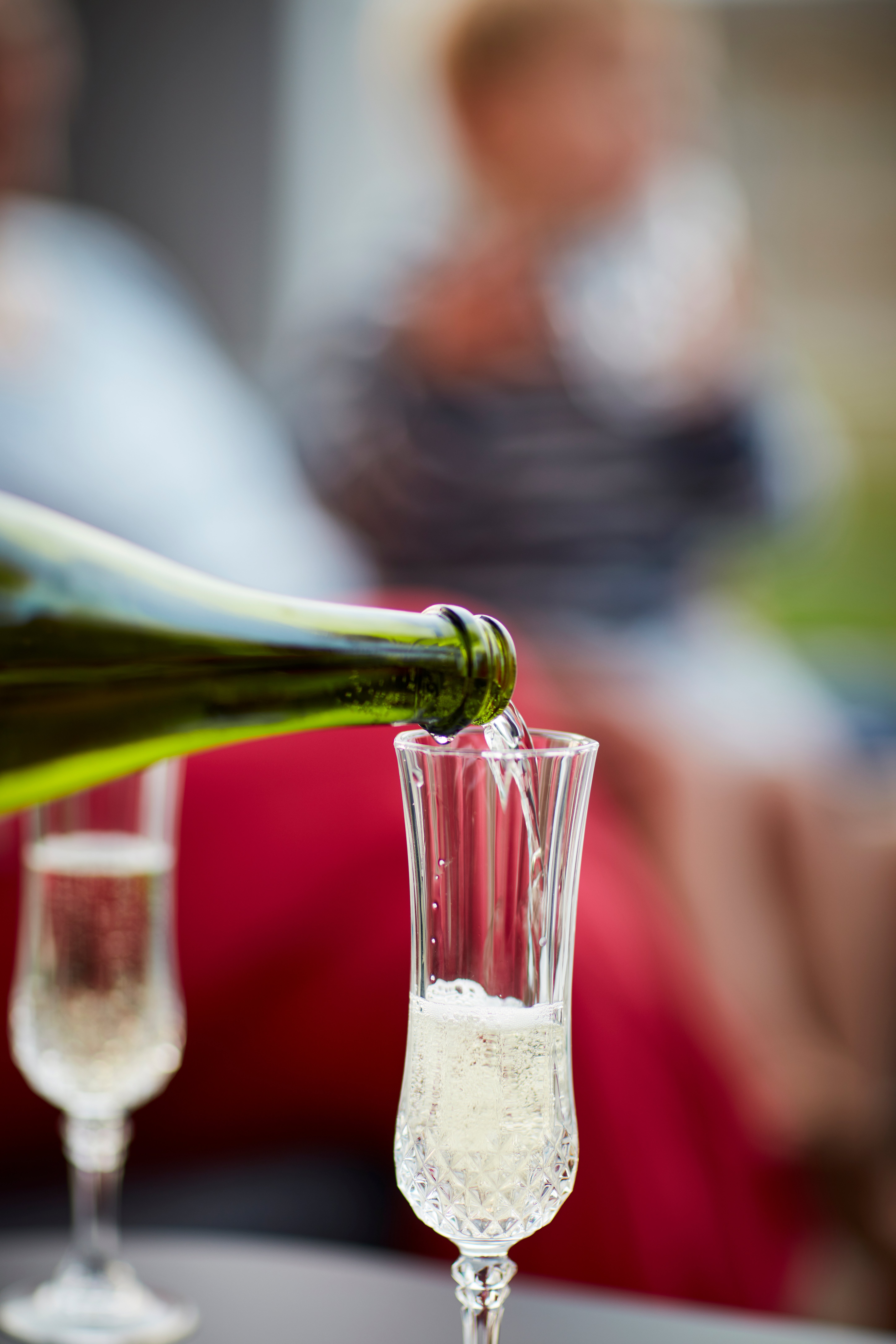   Gros plan sur du vin mousseux versé dans une flûte. À l'extérieur, des personnes sont rassemblées, floues au second plan.